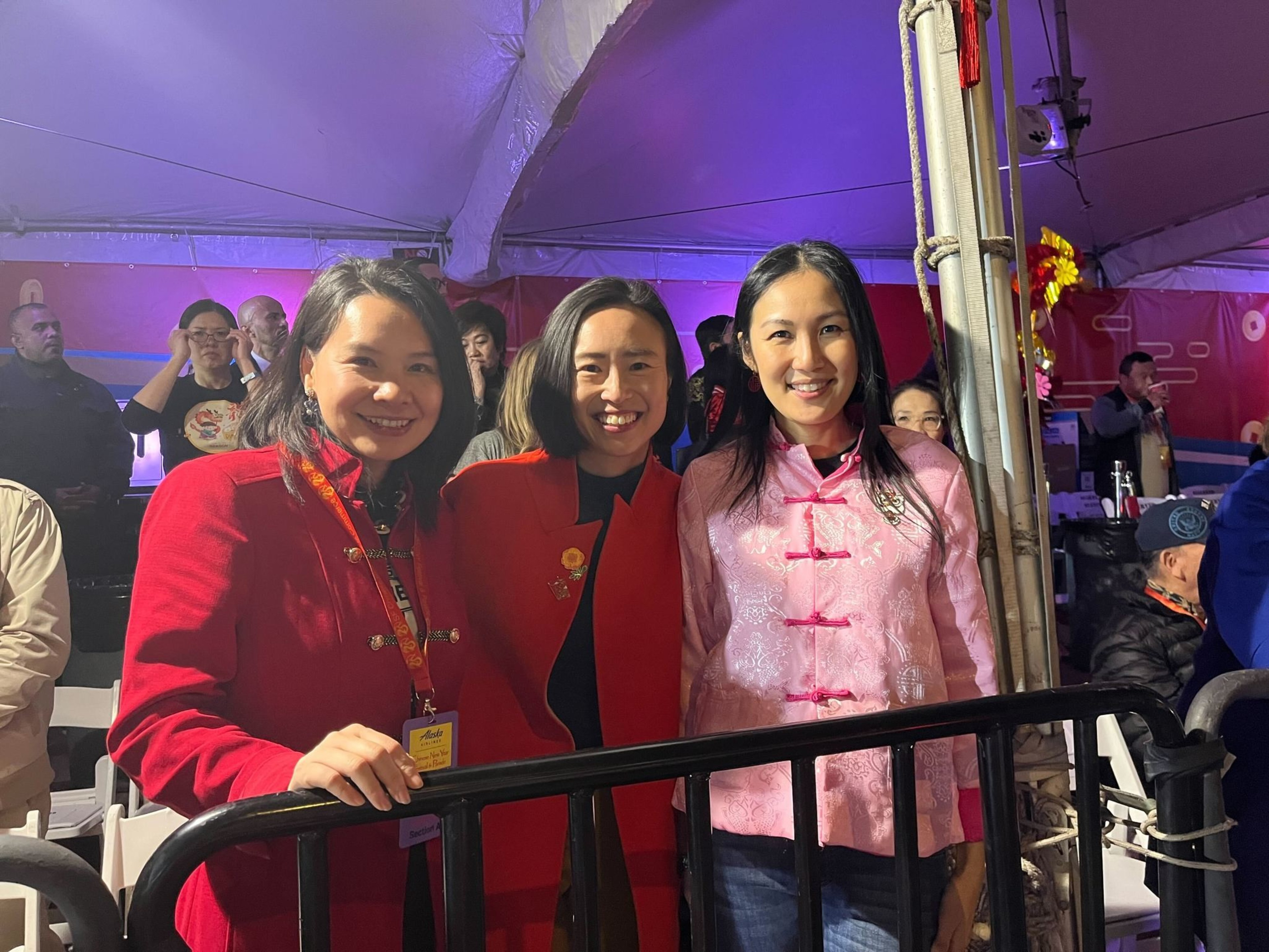 Three smiling women in traditional attire stand behind a railing against a festive backdrop.