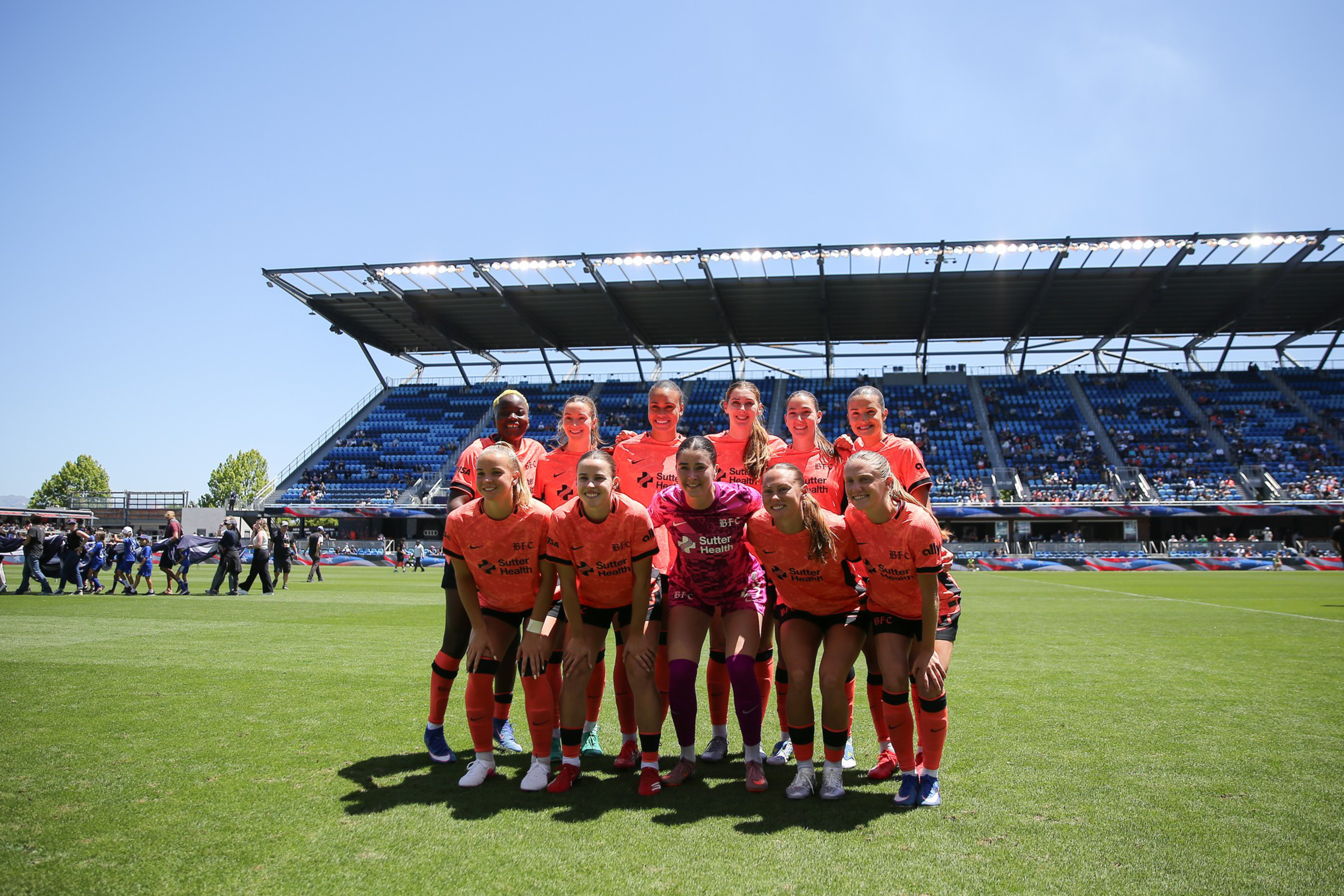 A women’s soccer team poses for a group photo on a sunny field with a stadium and spectators in the background.