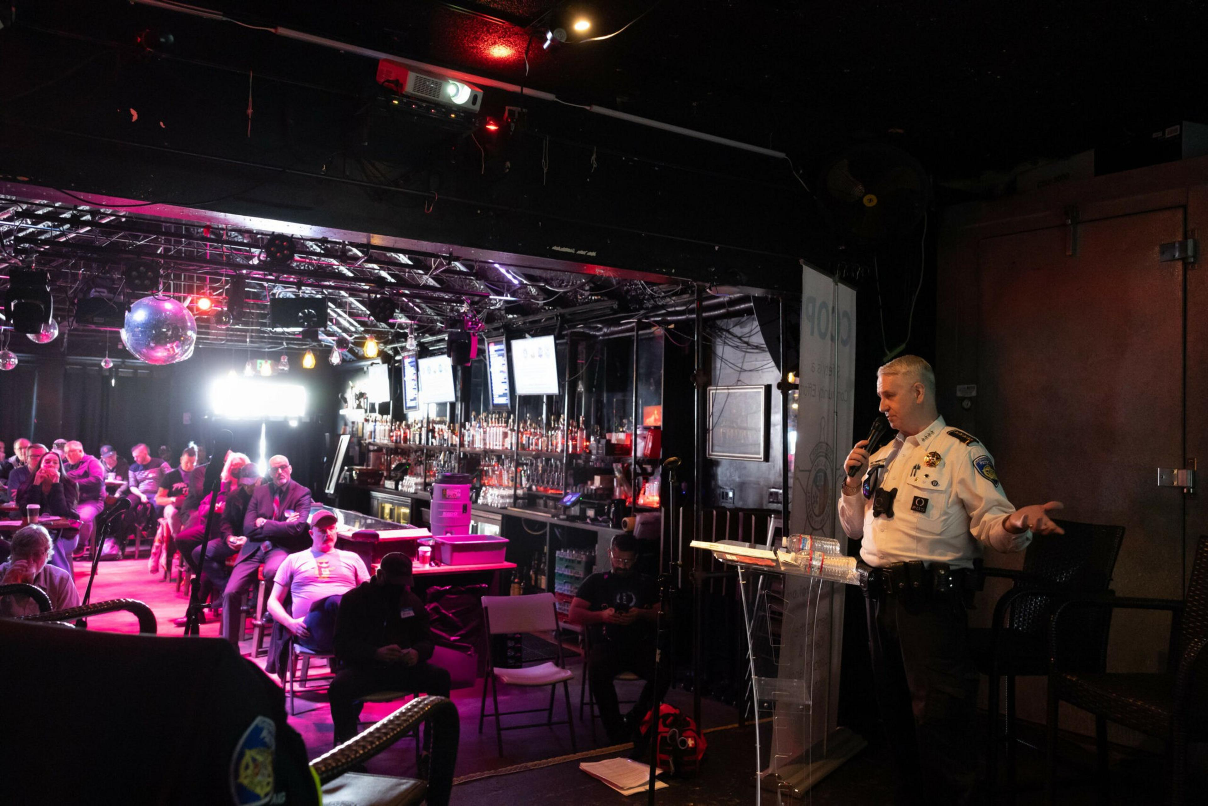A police officer speaks into a microphone in a dimly lit bar with seated audience members facing him and a disco ball hanging from the ceiling.