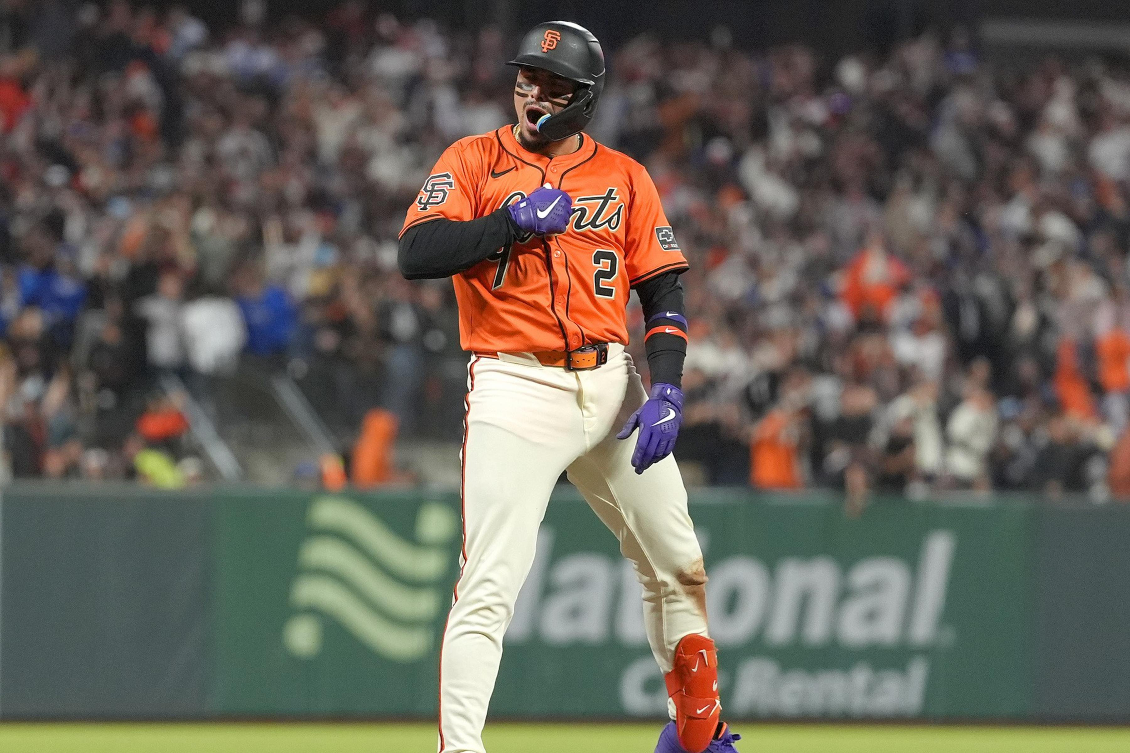 A baseball player in an orange jersey and white pants celebrates on a base. He wears a helmet, purple gloves, and bright orange protective gear amid a cheering crowd.