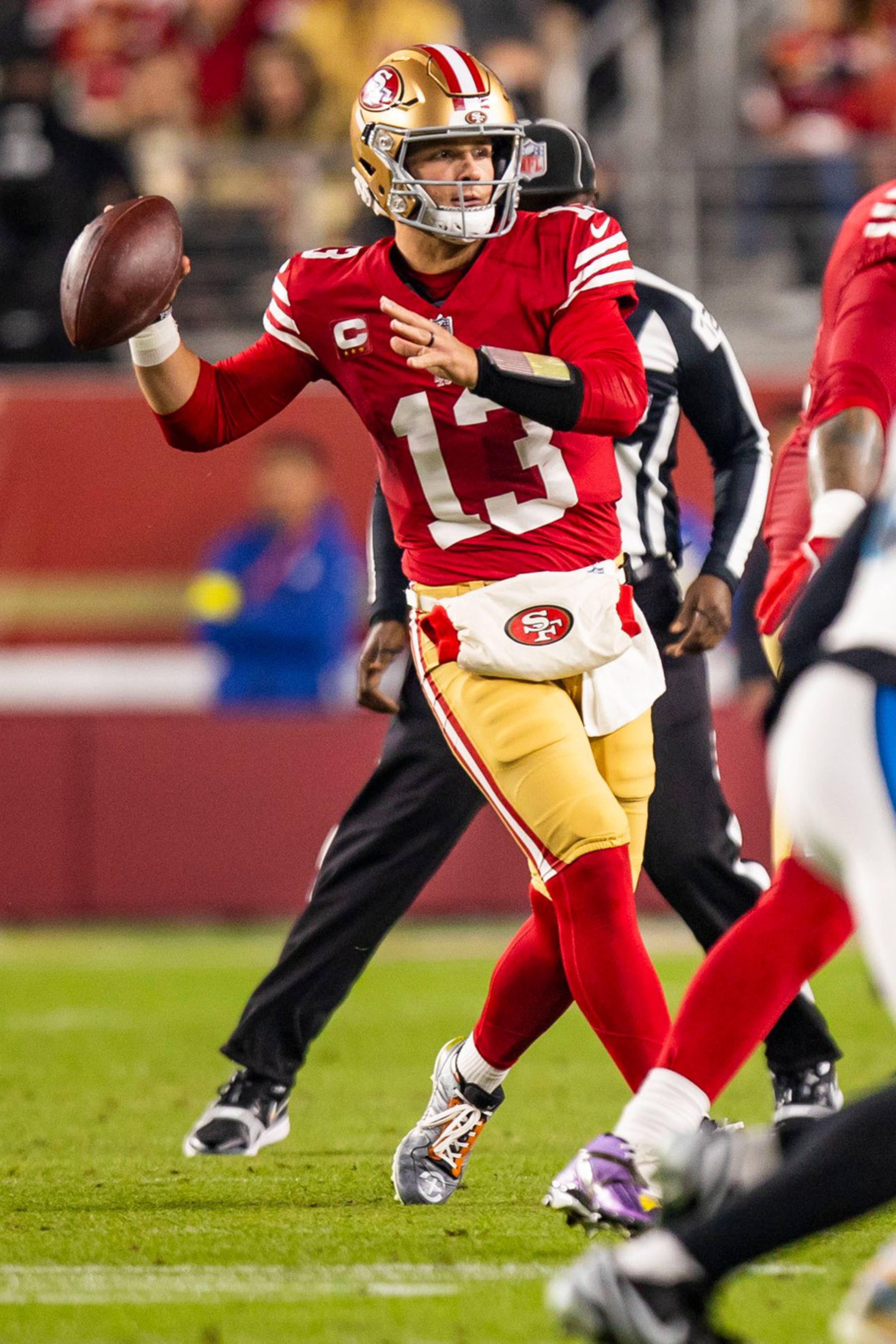 A quarterback in a red San Francisco 49ers uniform prepares to throw a football during a game, with teammates and opponents nearby.