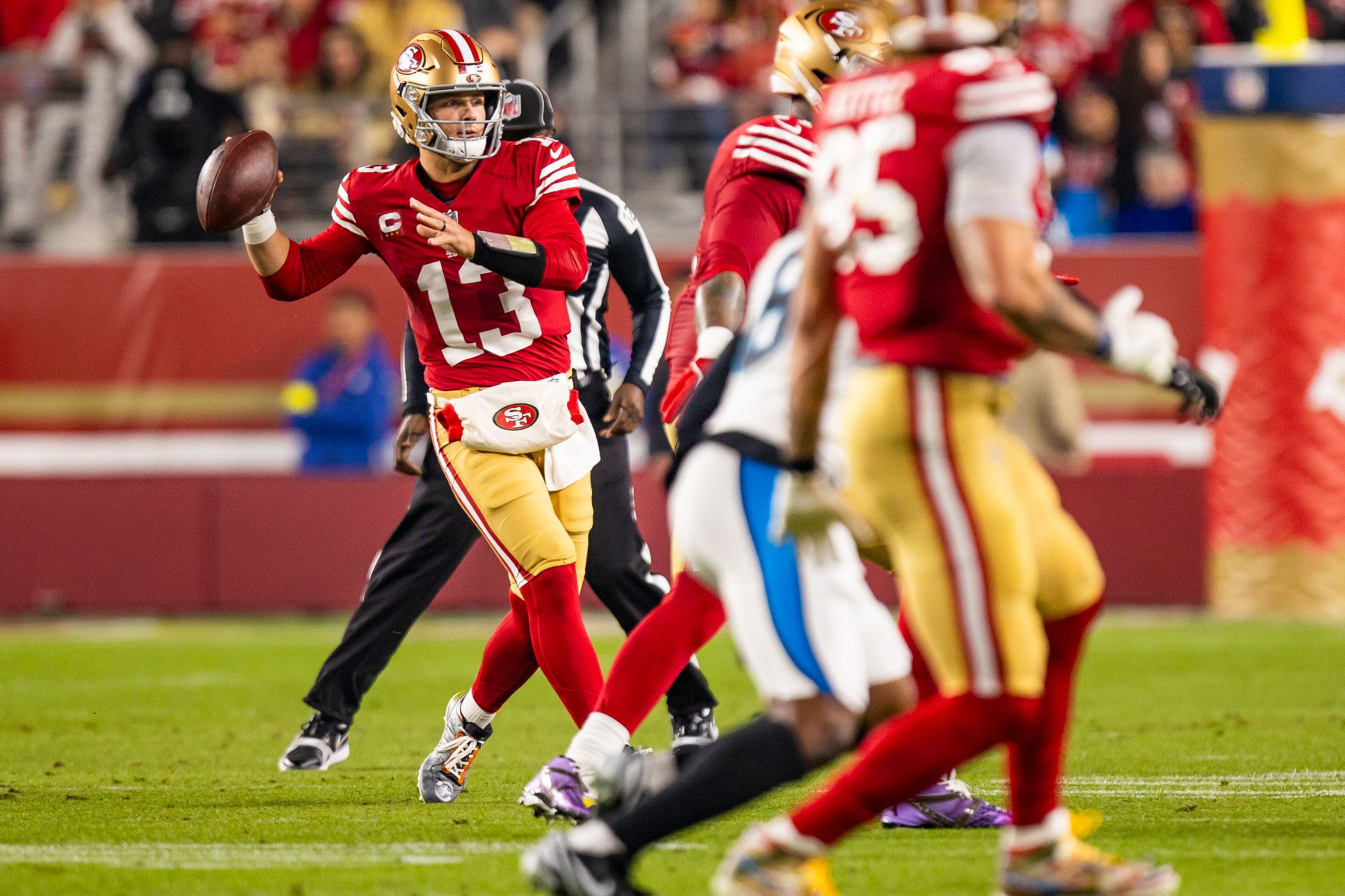 A quarterback in a red San Francisco 49ers uniform prepares to throw a football during a game, with teammates and opponents nearby.