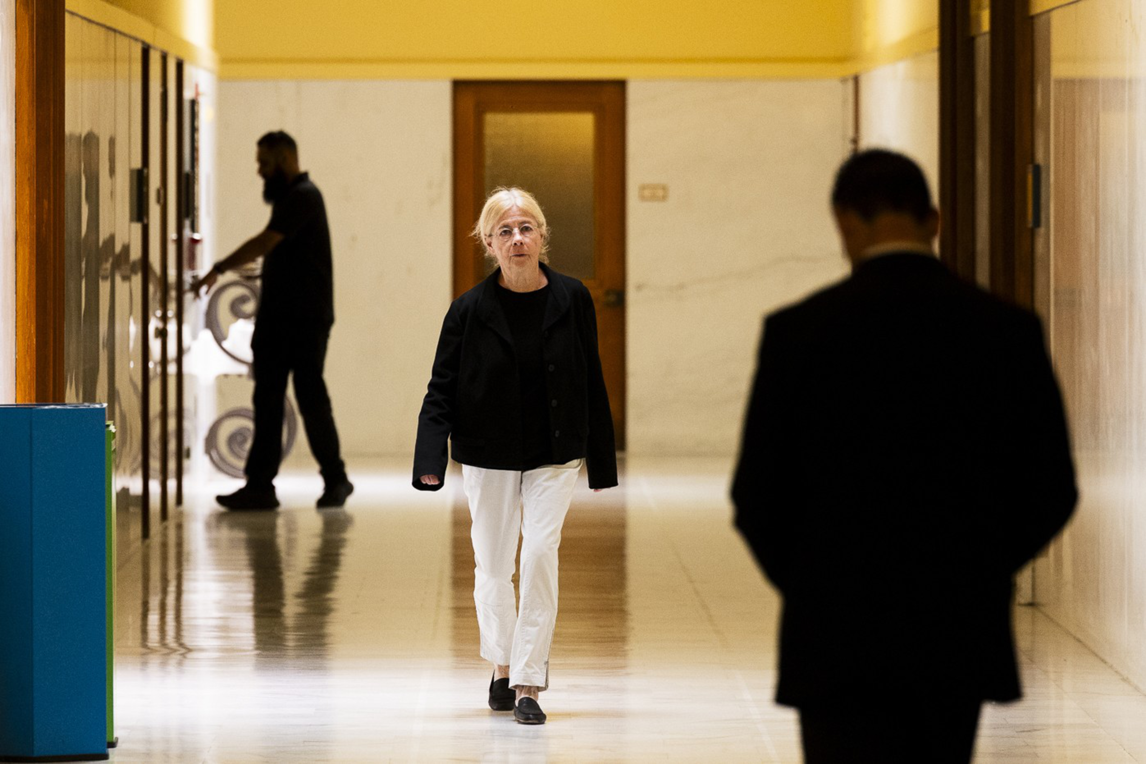 A person in white pants and a black jacket walks down a corridor, while another figure is silhouetted in the foreground. A third person stands to the side.