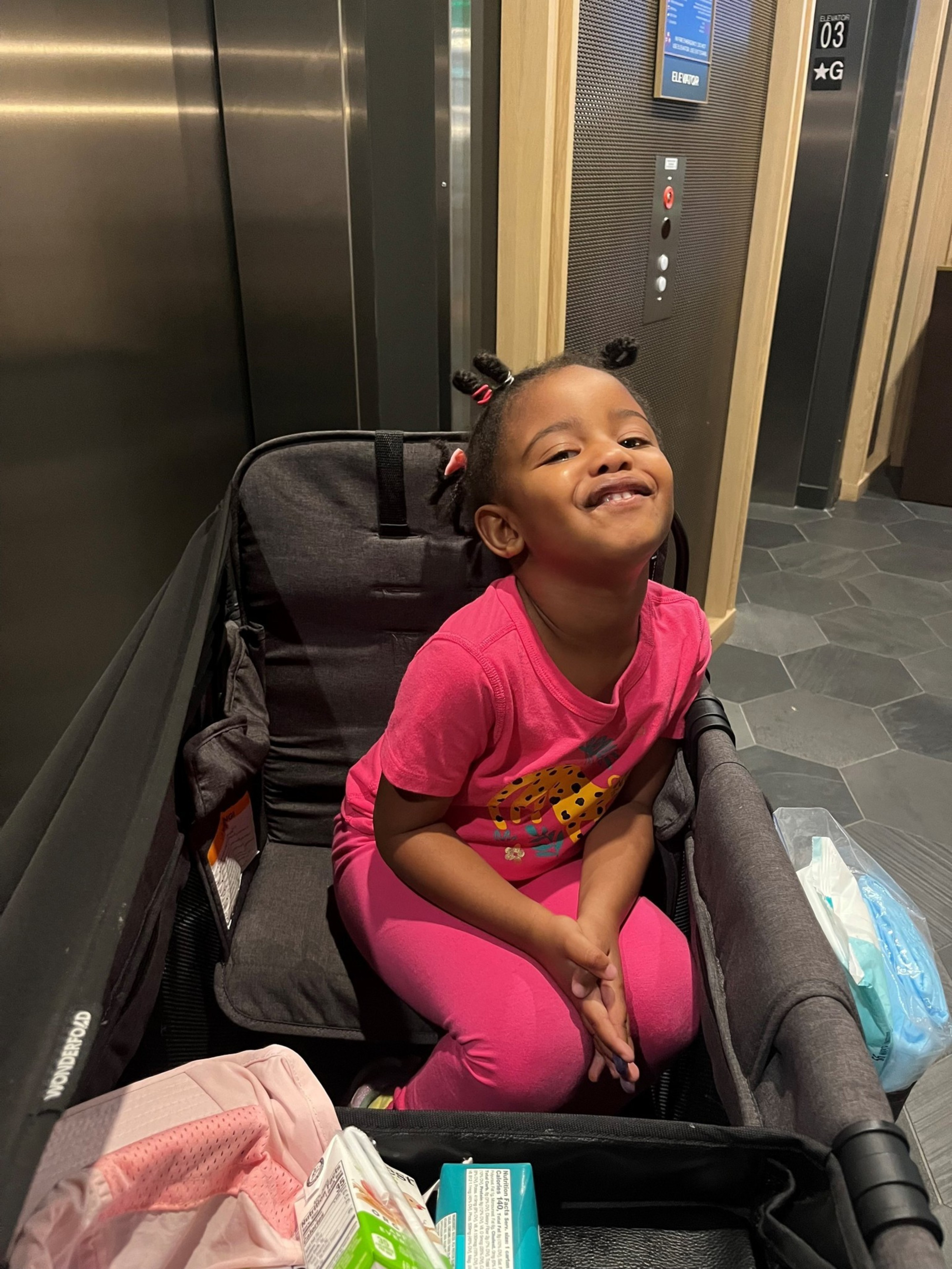 A girl with pink clothing and braided hair sits in a stroller, smiling. Around her are various items like packages and clothing. The background features an elevator.