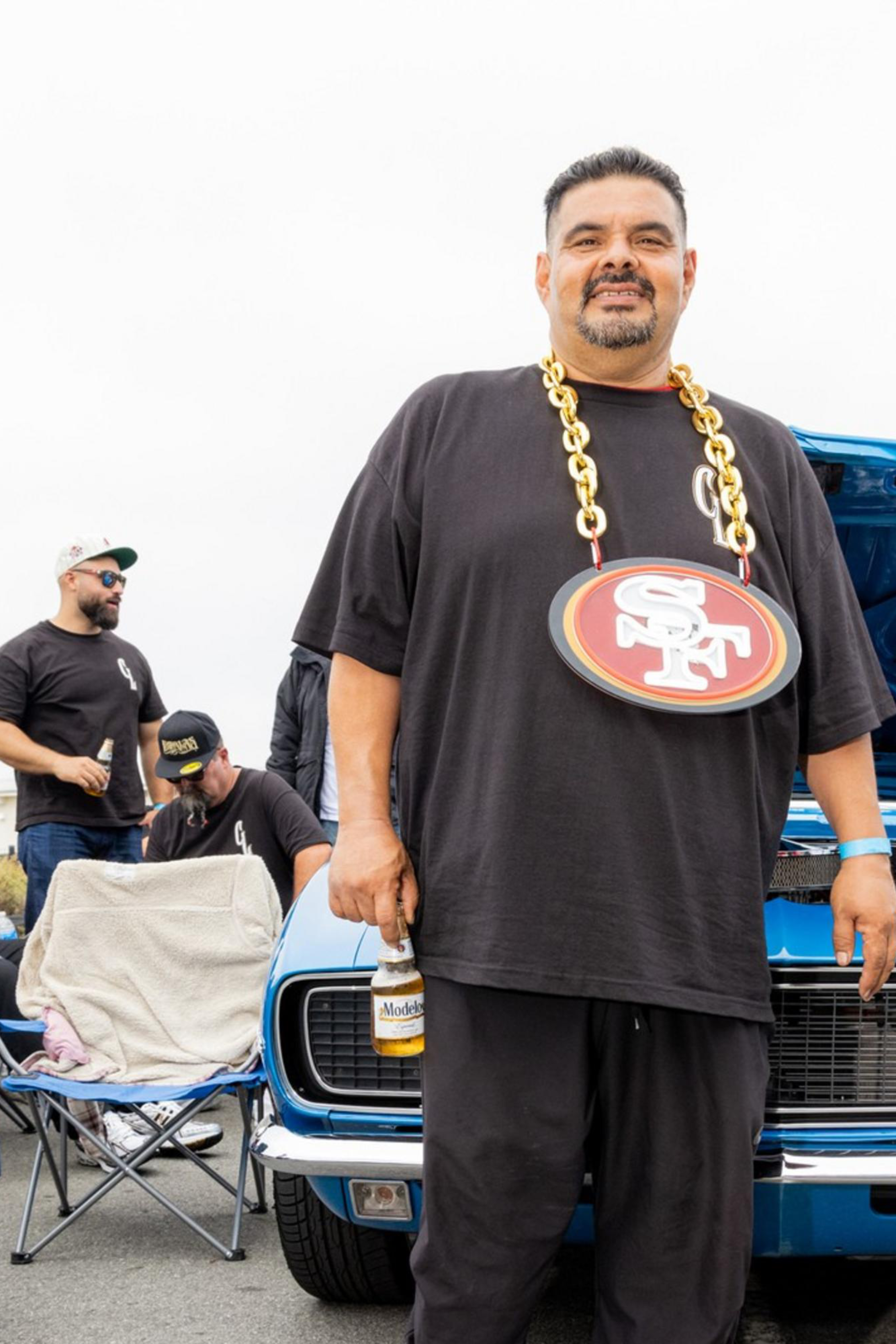 A man stands in front of a blue car with its hood open, wearing a large chain with a football team logo. Other people relax in chairs around a red convertible and another vehicle.