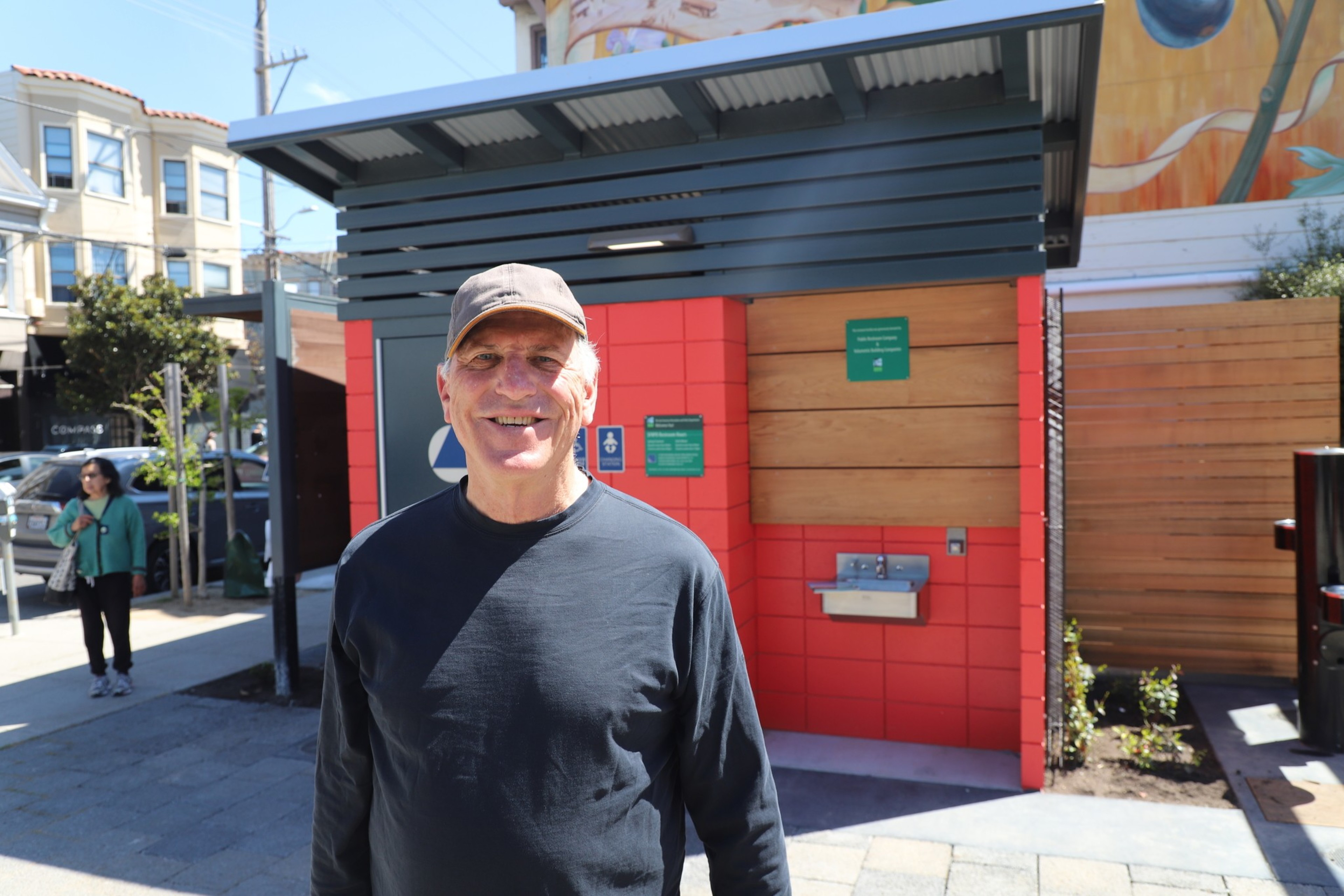 A man with white hair wearing a dark long-sleeve T-shirt and light-colored baseball cap smiles in front of a public restroom.