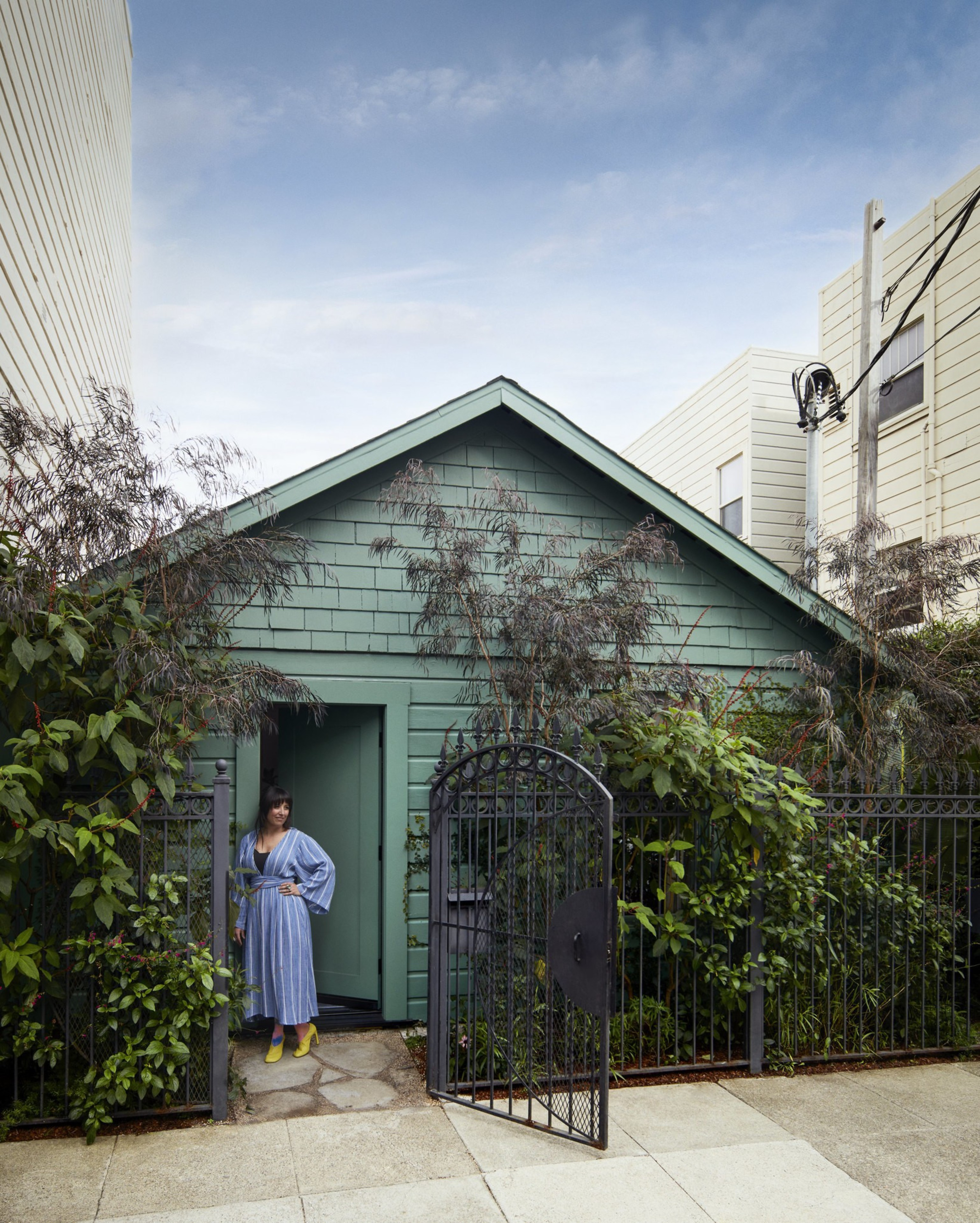 A woman in a blue dress and yellow shoes stands in the doorway of a small green house surrounded by tall plants and an open black metal gate.