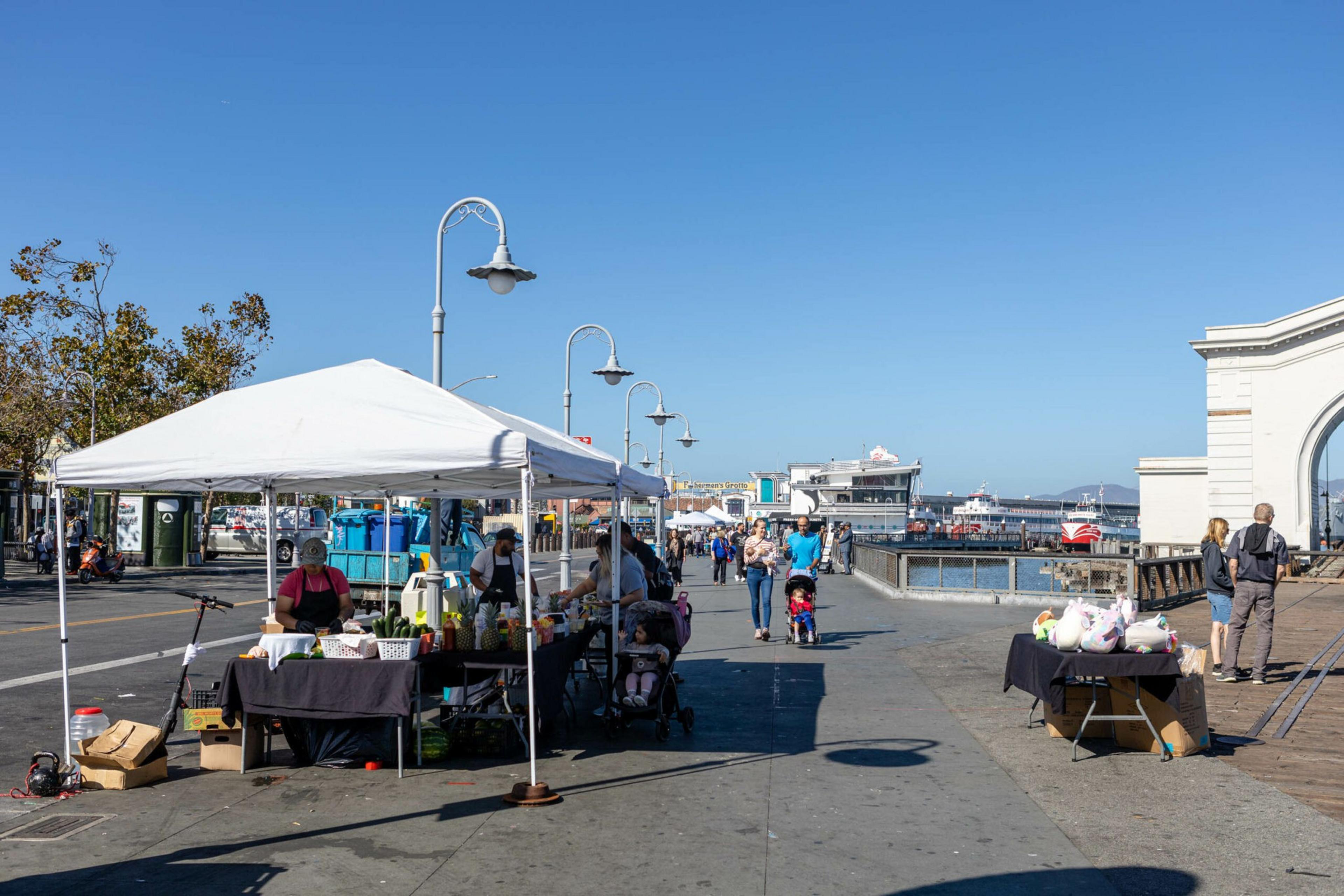 Street market stalls with vendors and shoppers line a sunny waterfront promenade, with boats docked and clear blue skies overhead.