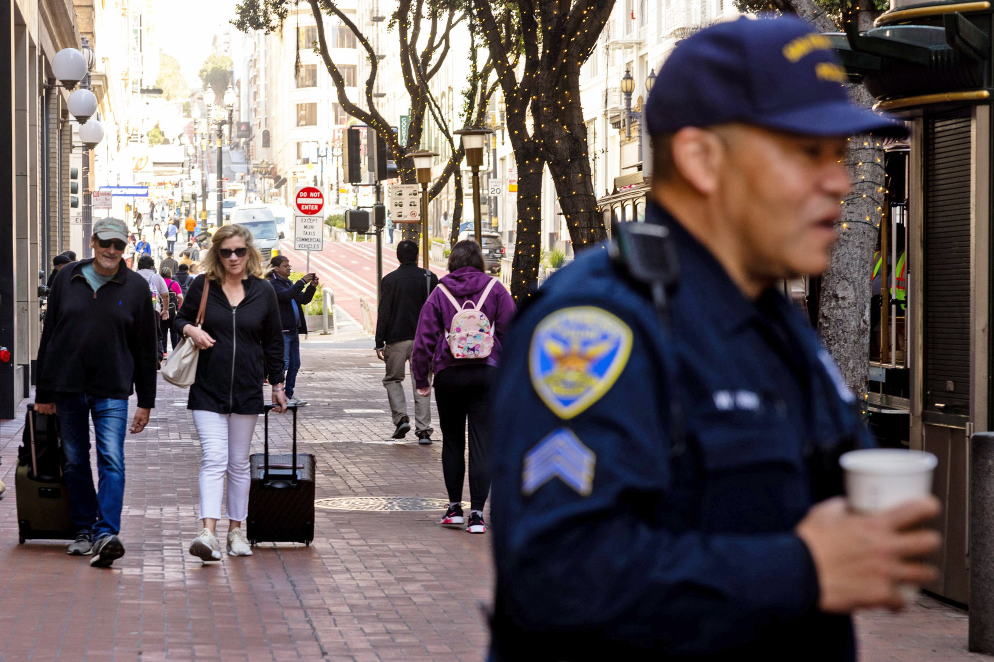 A busy city street scene featuring a police officer in the foreground, people walking, and some carrying luggage, with buildings and trees adorned with lights in the background.