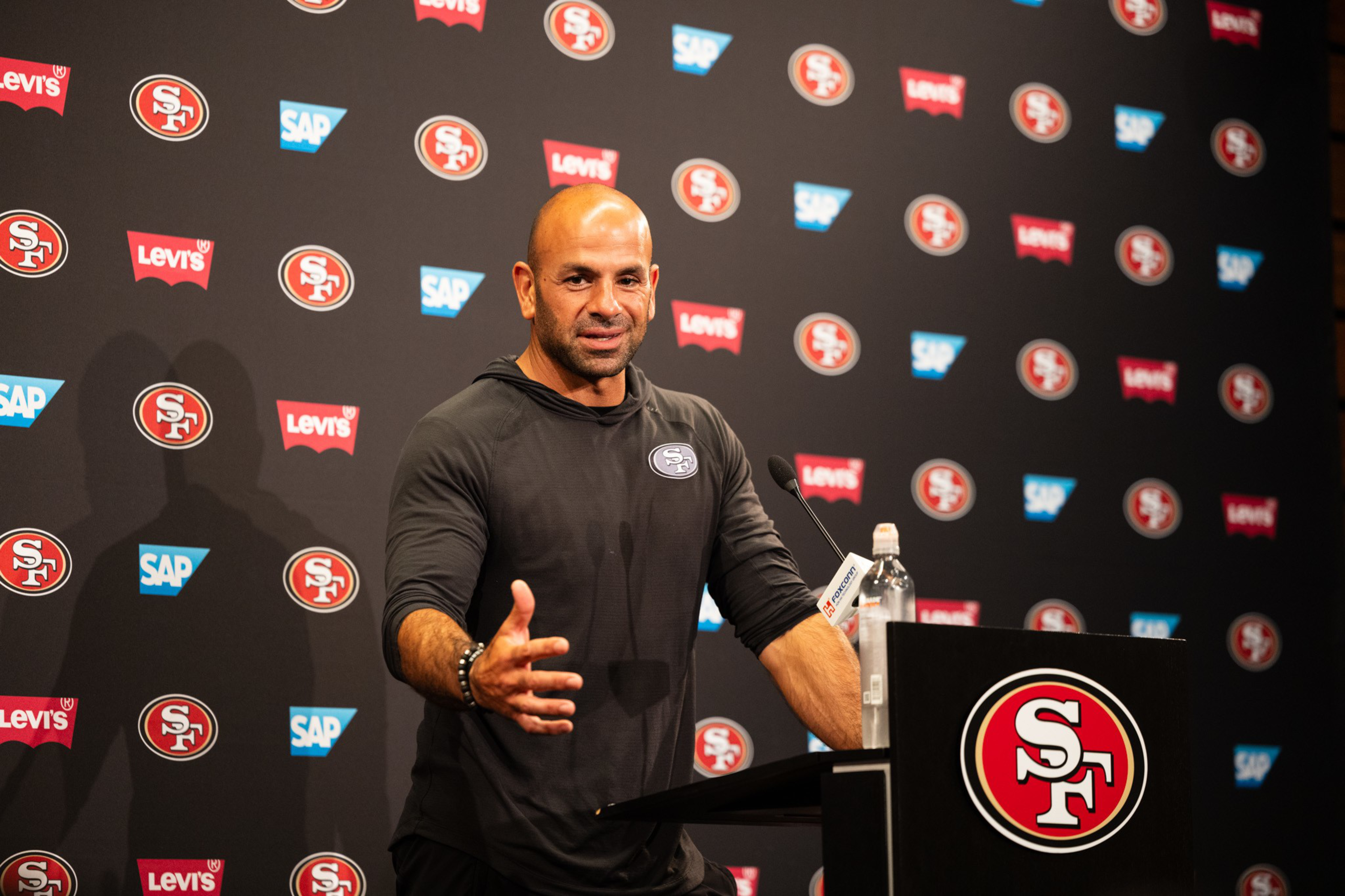 A man stands at a podium with an &quot;SF&quot; logo, addressing media. He gestures with his hand. The backdrop displays logos of the 49ers, SAP, and Levi's.
