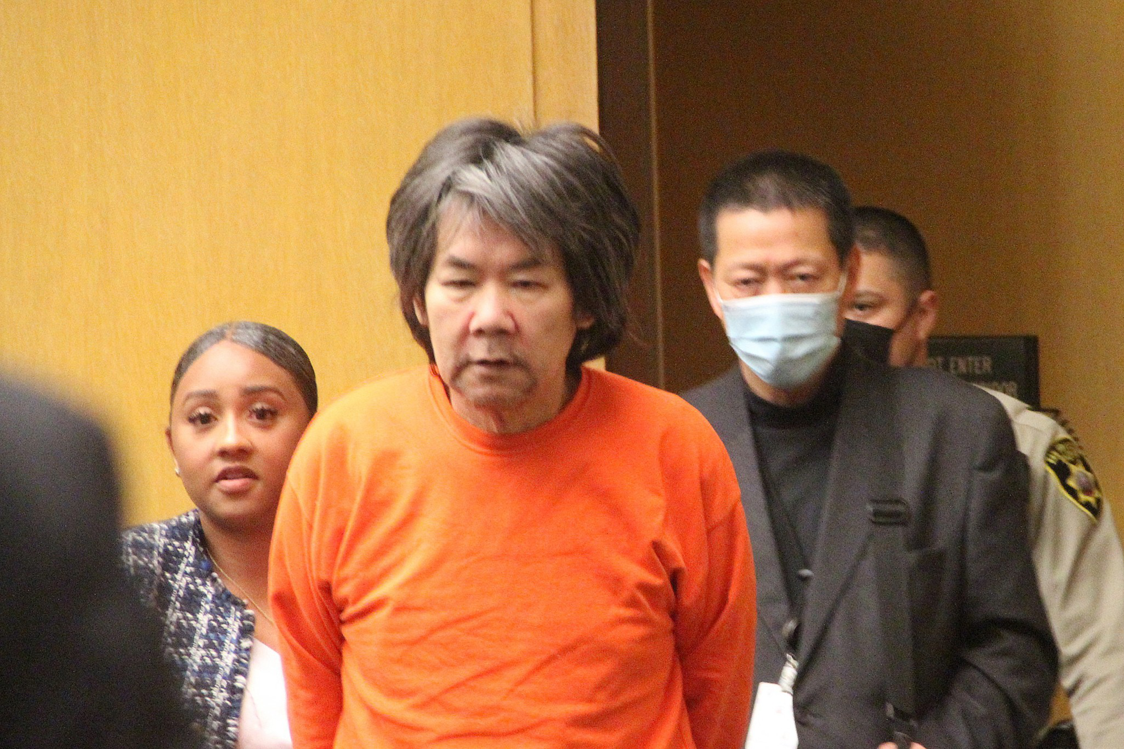 A man in an orange shirt is being escorted by a woman, a masked man, and a uniformed officer in a hallway with a wooden wall.