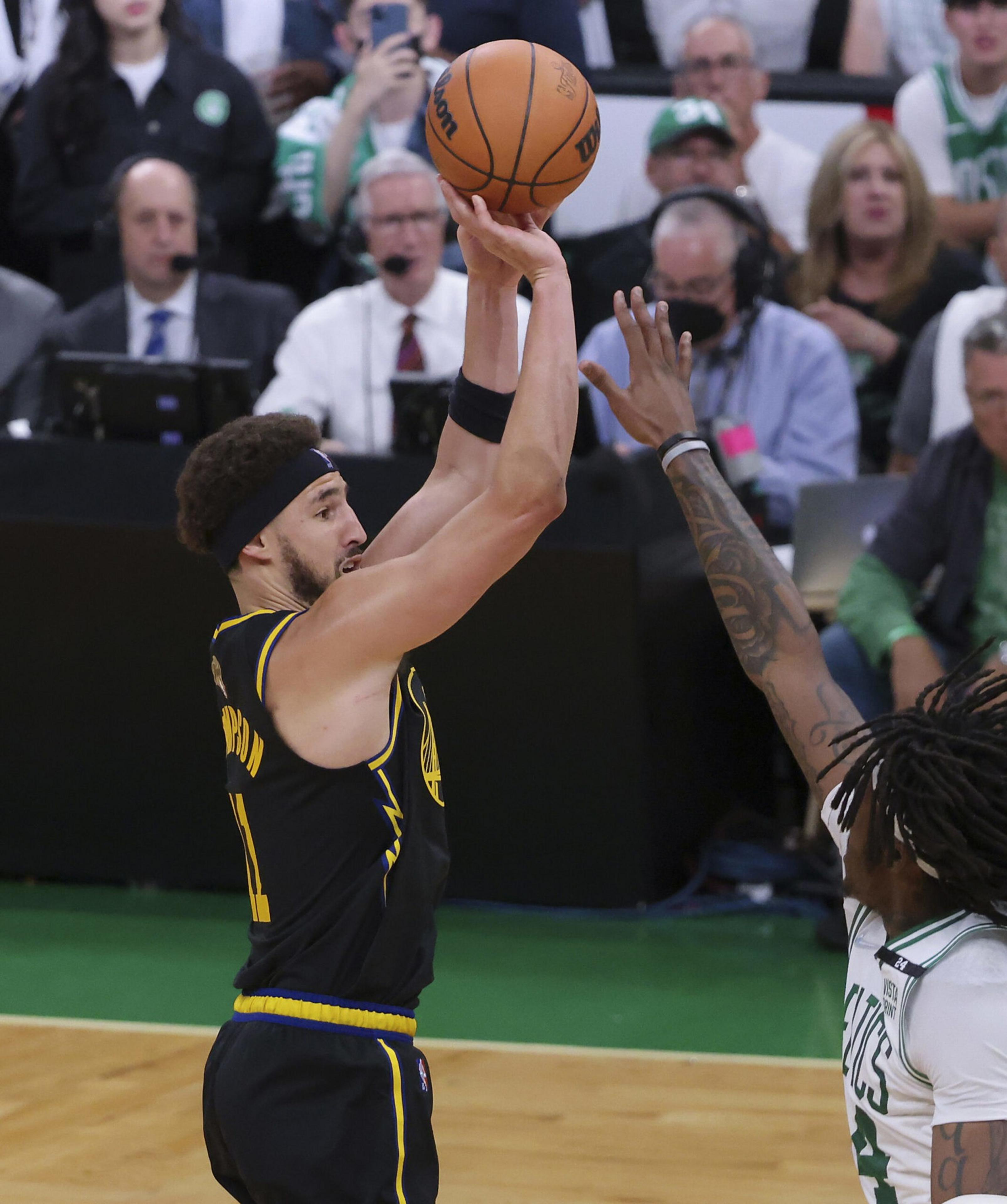 A basketball player in a black and yellow uniform is shooting the ball while a defender in a white and green uniform attempts to block the shot.