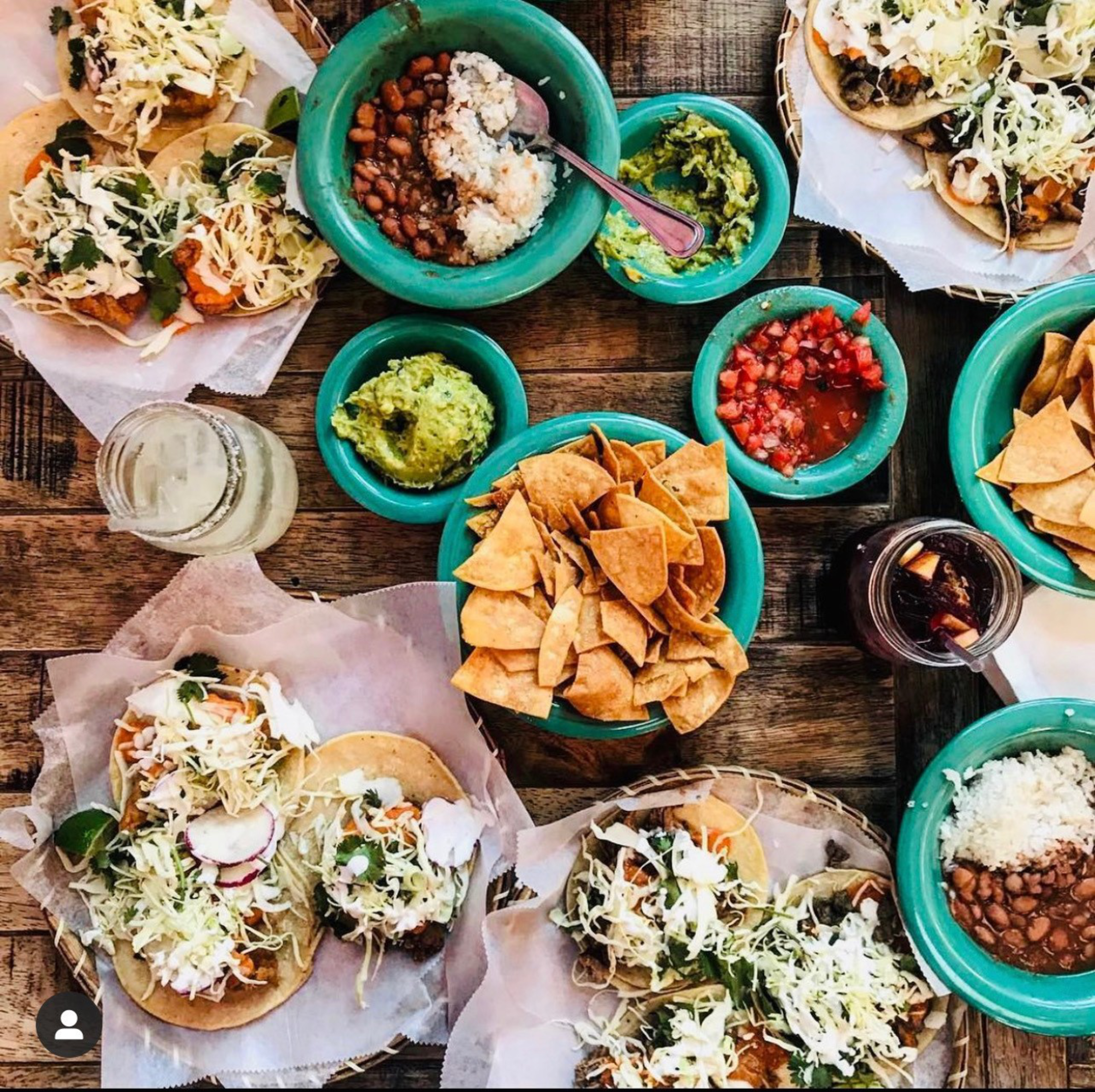 Tacos with shredded cabbage, radishes, and sour cream surround bowls of chips, guacamole, salsa, beans, rice, and two drinks on a wooden table.
