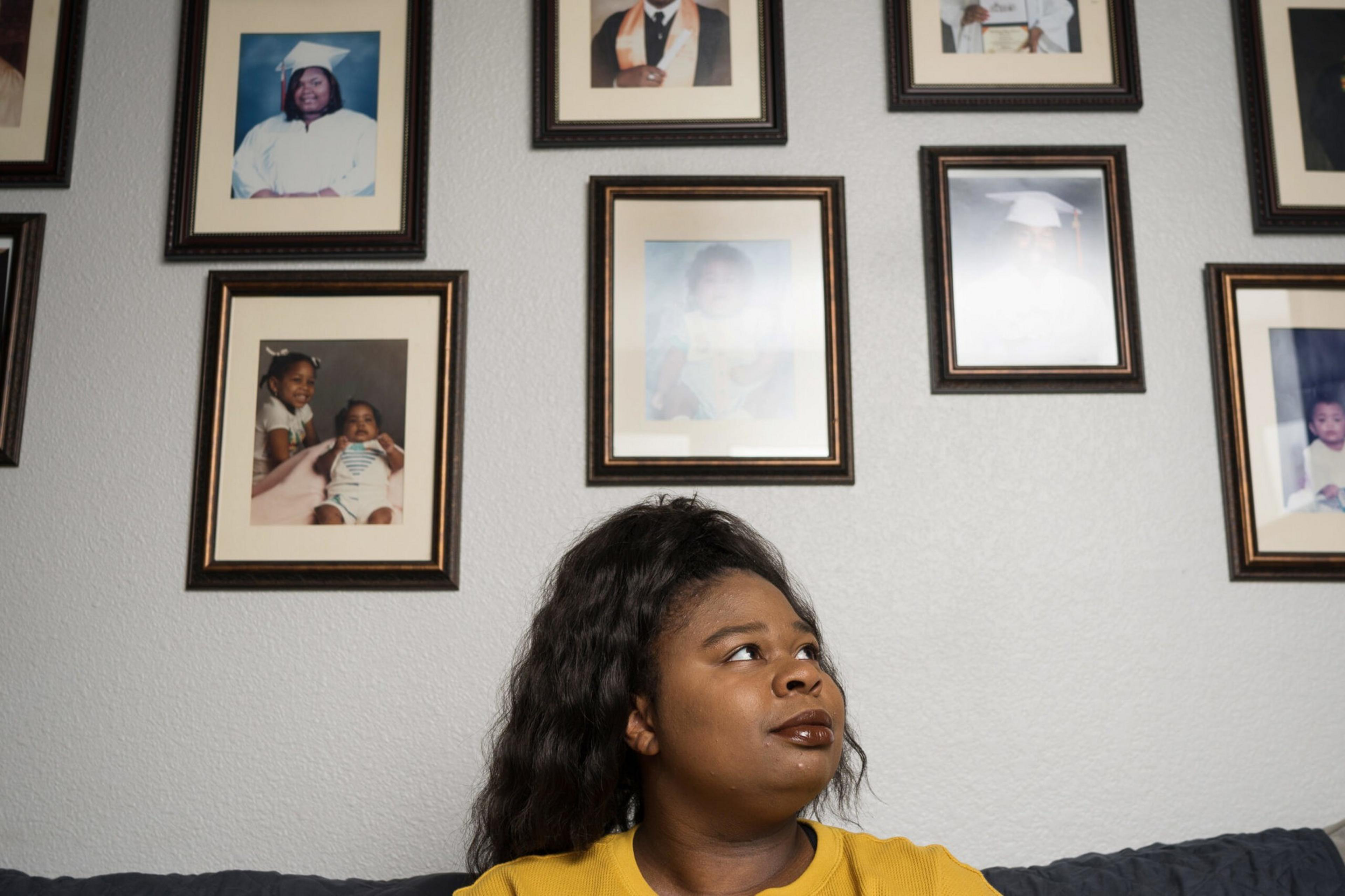 A woman with long dark hair wearing a yellow top sits on a couch, looking up thoughtfully at framed family photos on the wall behind her.