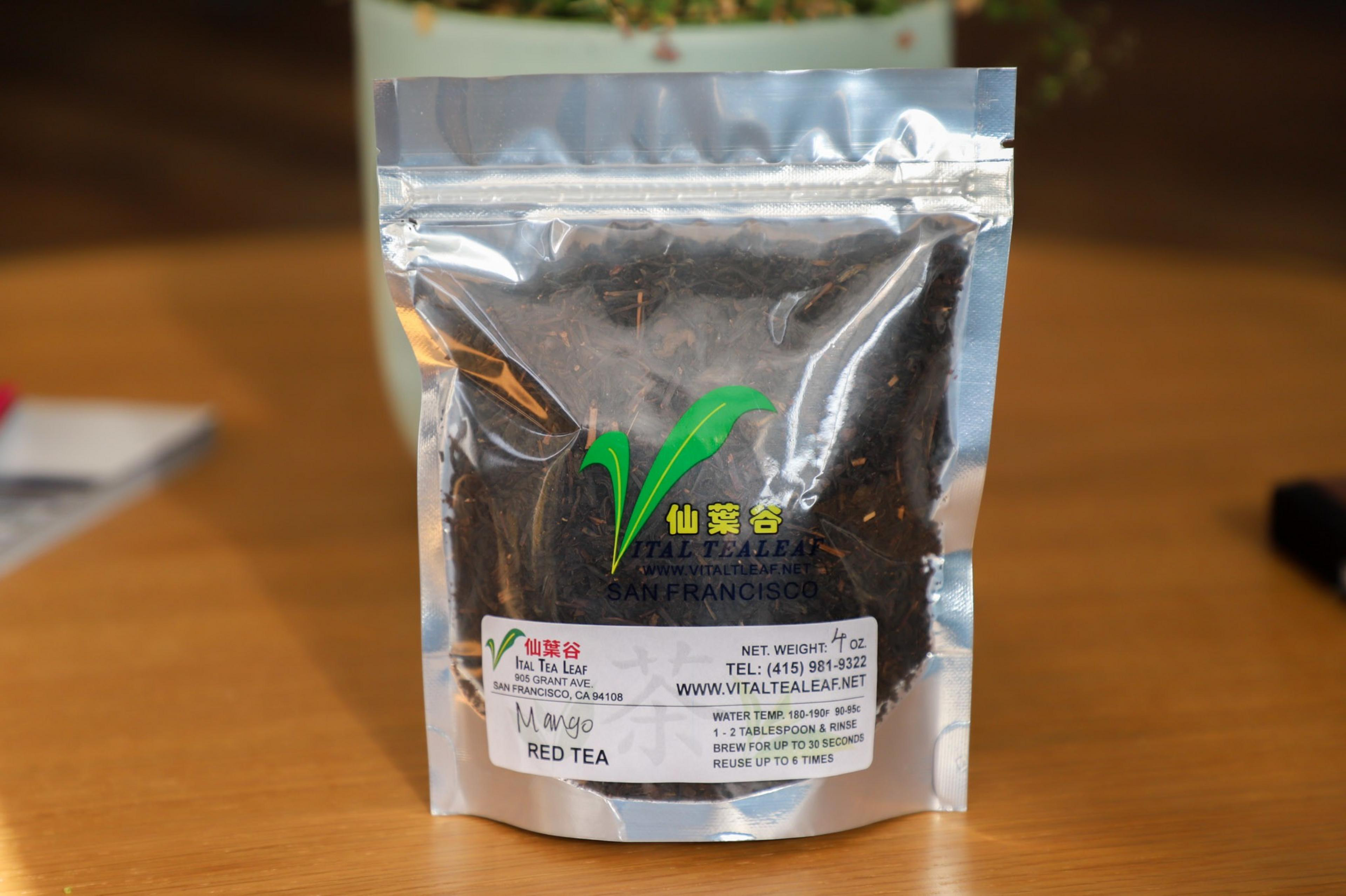 A silver, see-through bag of tea rests on a table with red tea inside it and a label that says mango red tea.