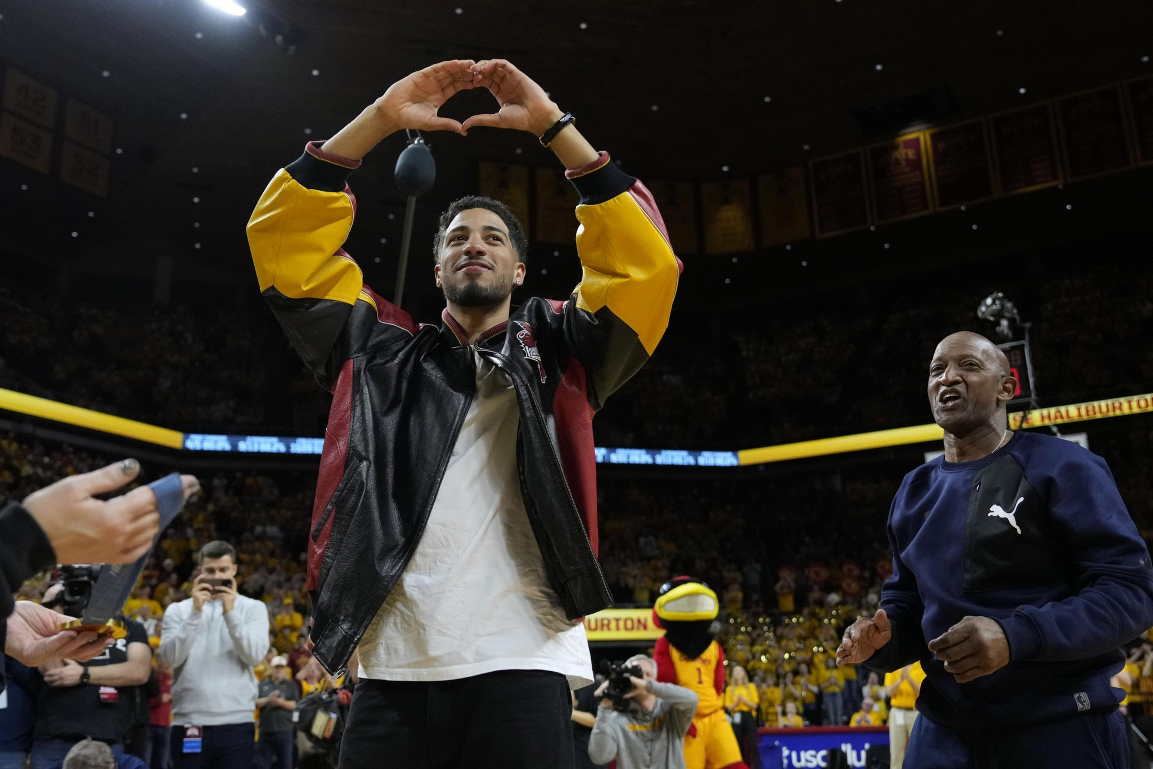 A person in a sports arena, wearing a colorful jacket, makes a heart shape with their hands. A man in a tracksuit stands nearby, and a mascot is visible behind them.
