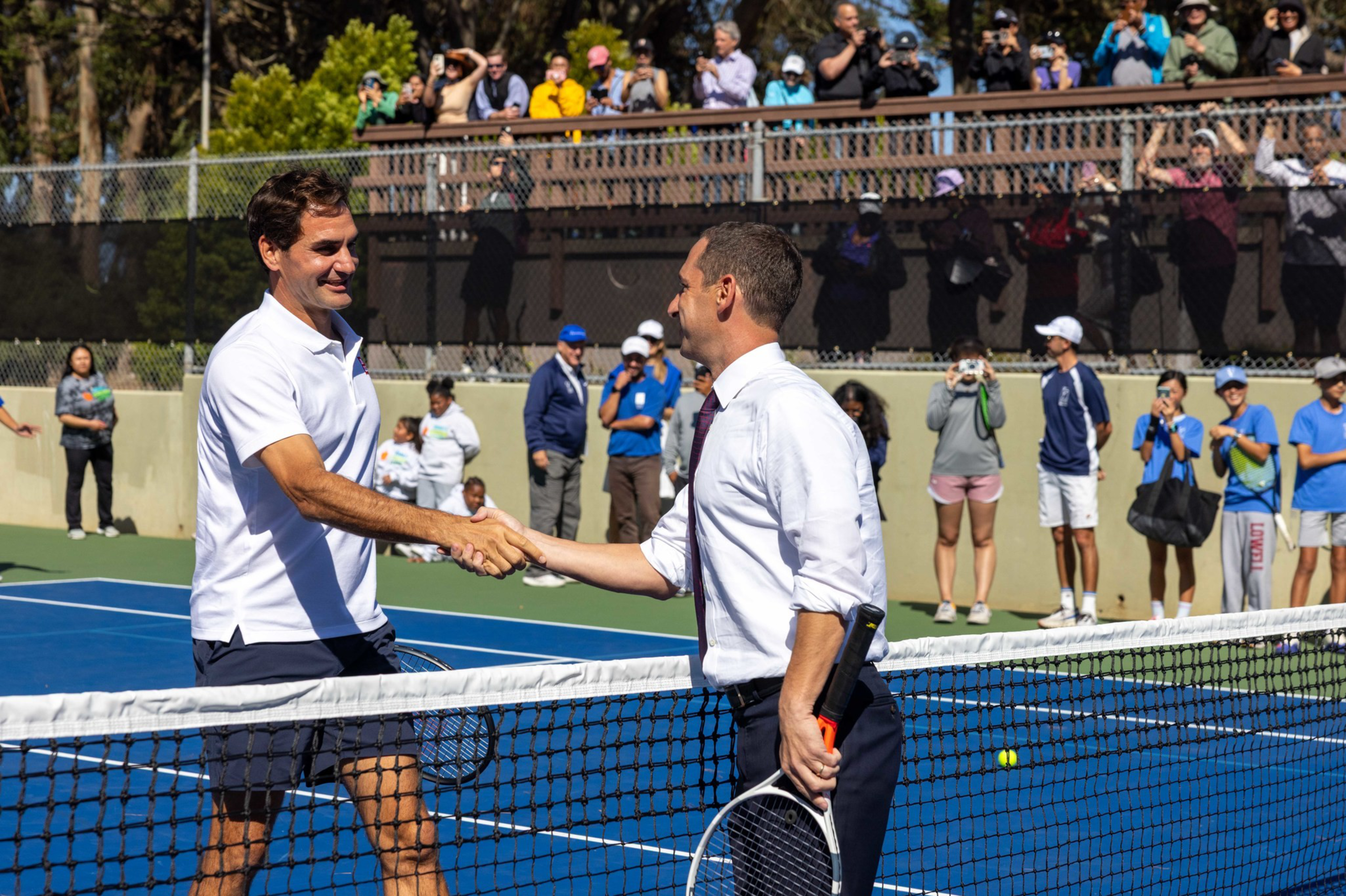 Two men shaking hands across a tennis net on a blue court, one in a white polo and shorts, the other in a white shirt and tie, with spectators in the background.