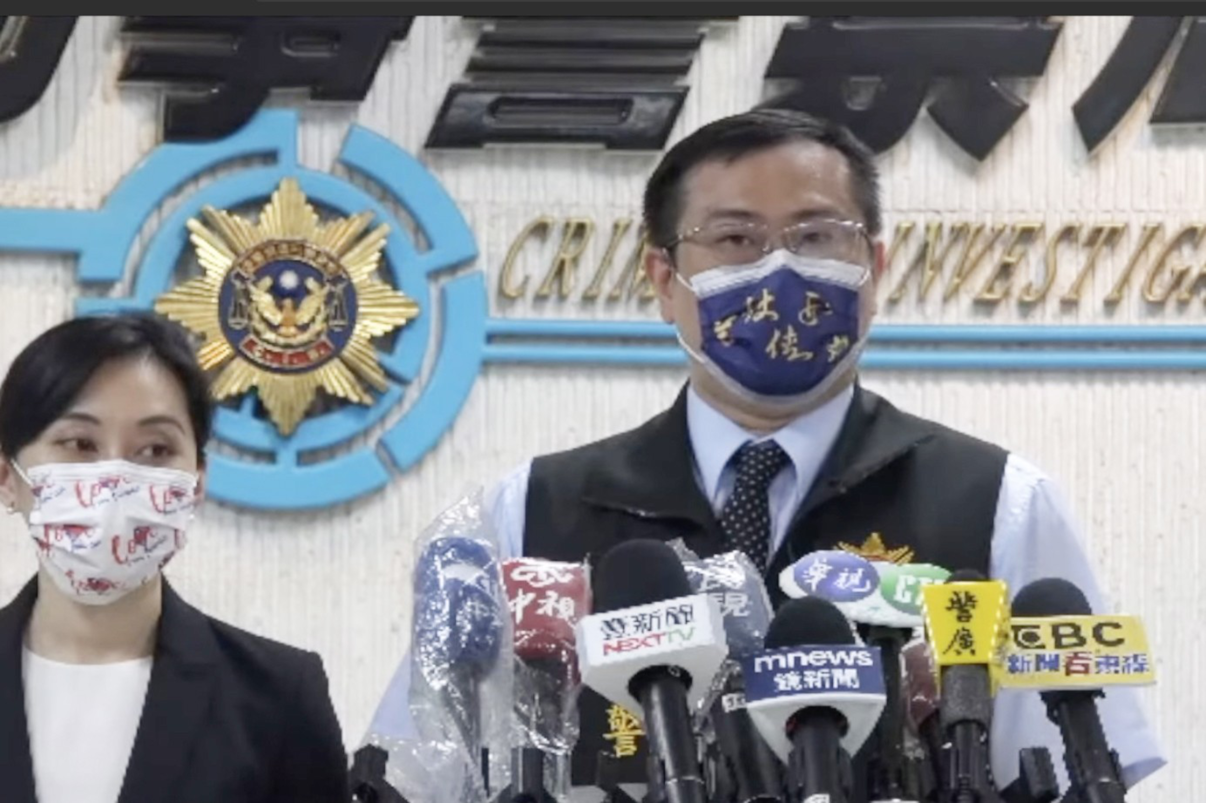 A man and a woman wearing face masks stand behind microphones at a press conference in front of a Crime Investigation logo.