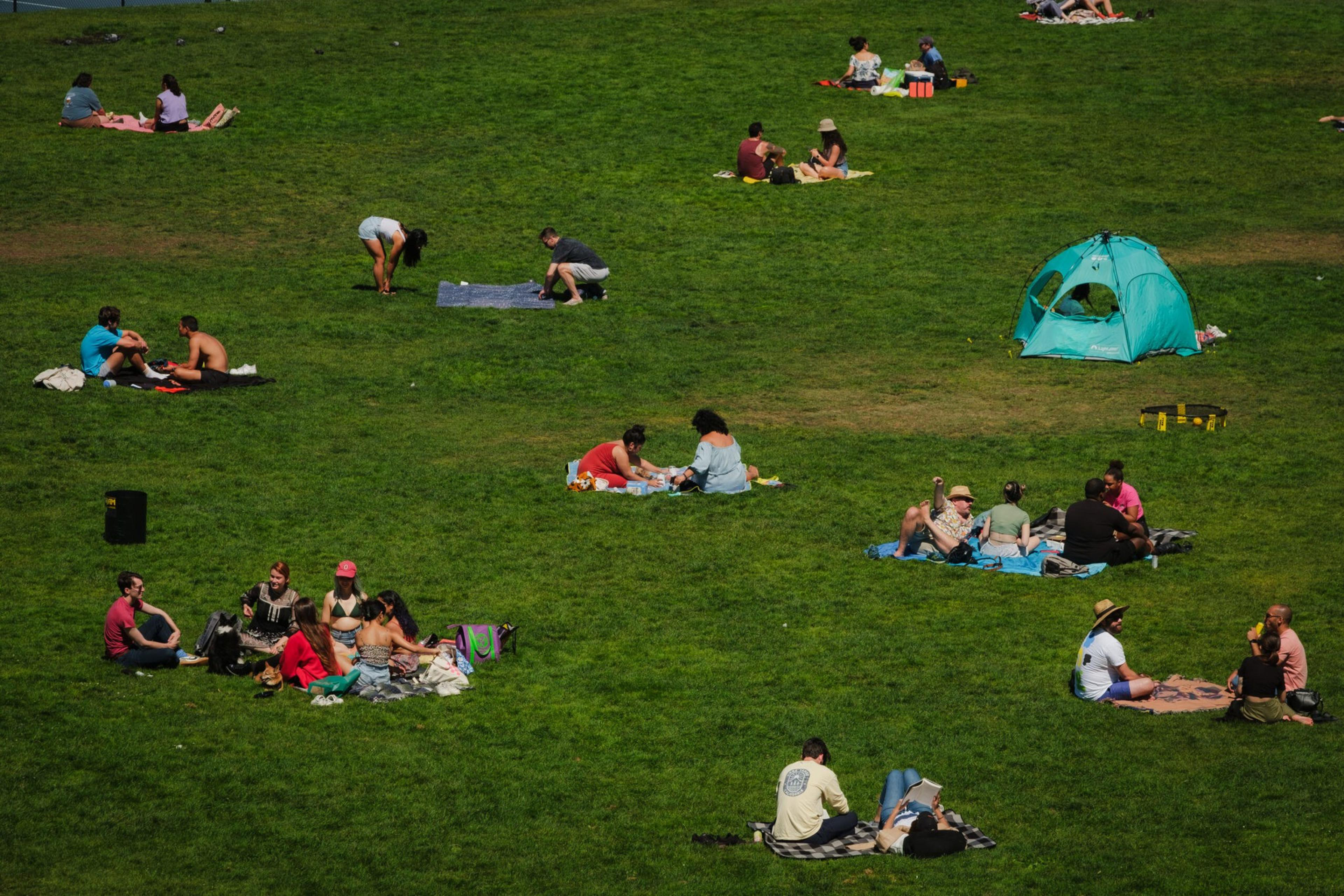 People are sitting or lying on blankets on a large grassy field, some chatting or relaxing, with a small blue tent set up on the right side.