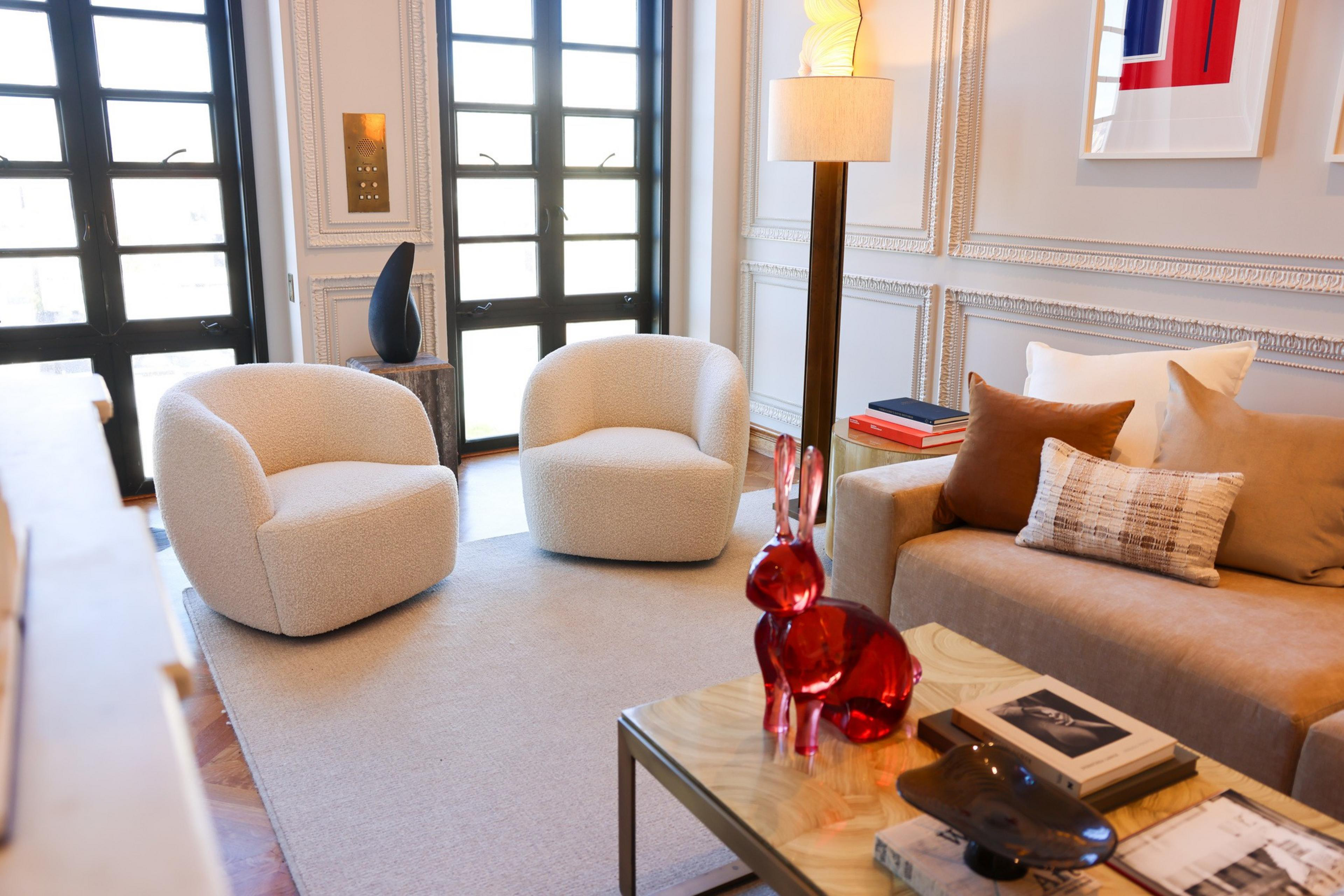 This room features two rounded beige chairs, a beige sofa with assorted pillows, a glass table with a red rabbit figurine, and a standing lamp.