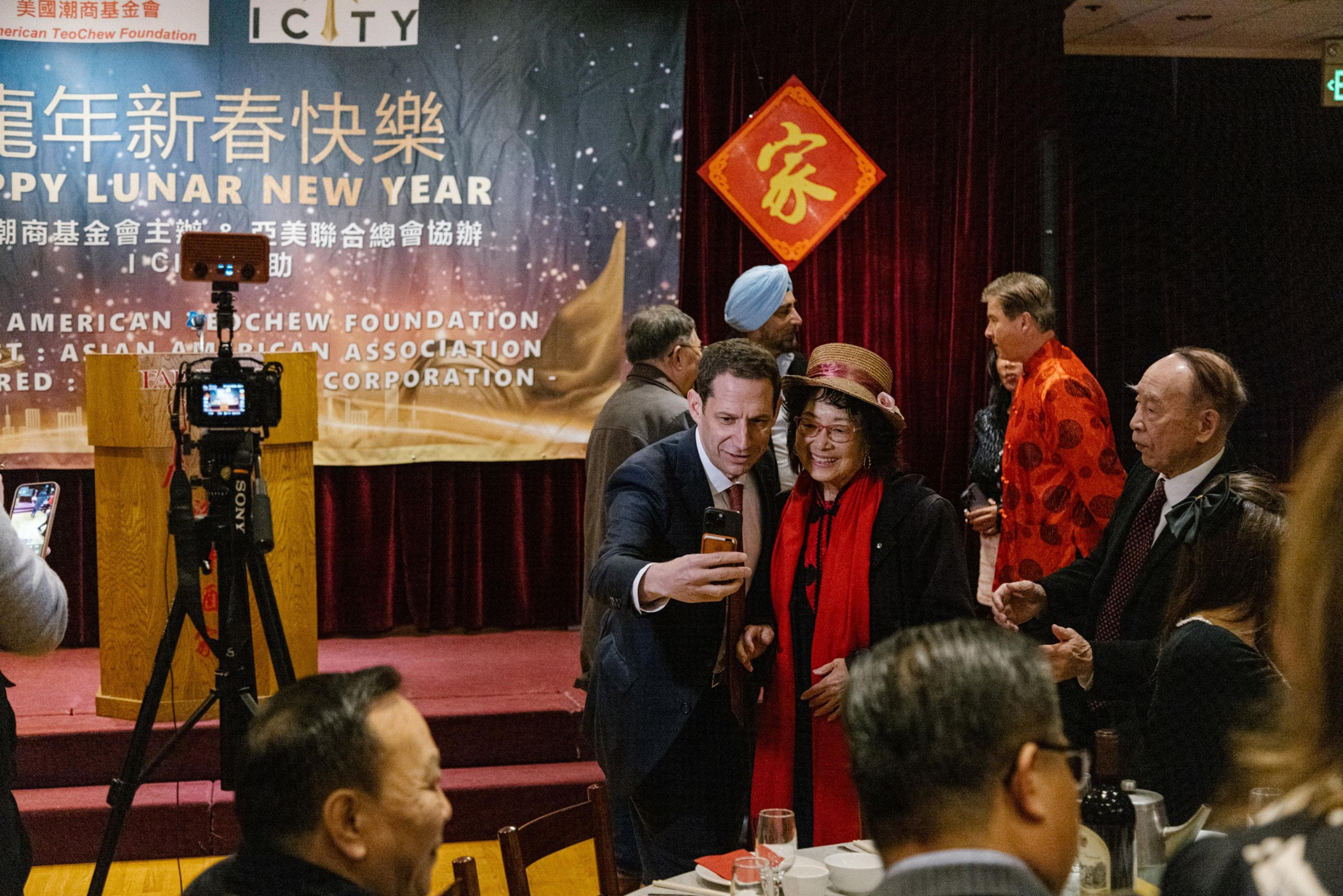 A group of people gather at a Lunar New Year event. Two people are posing for a selfie in the foreground. A festive banner and decorations are visible.