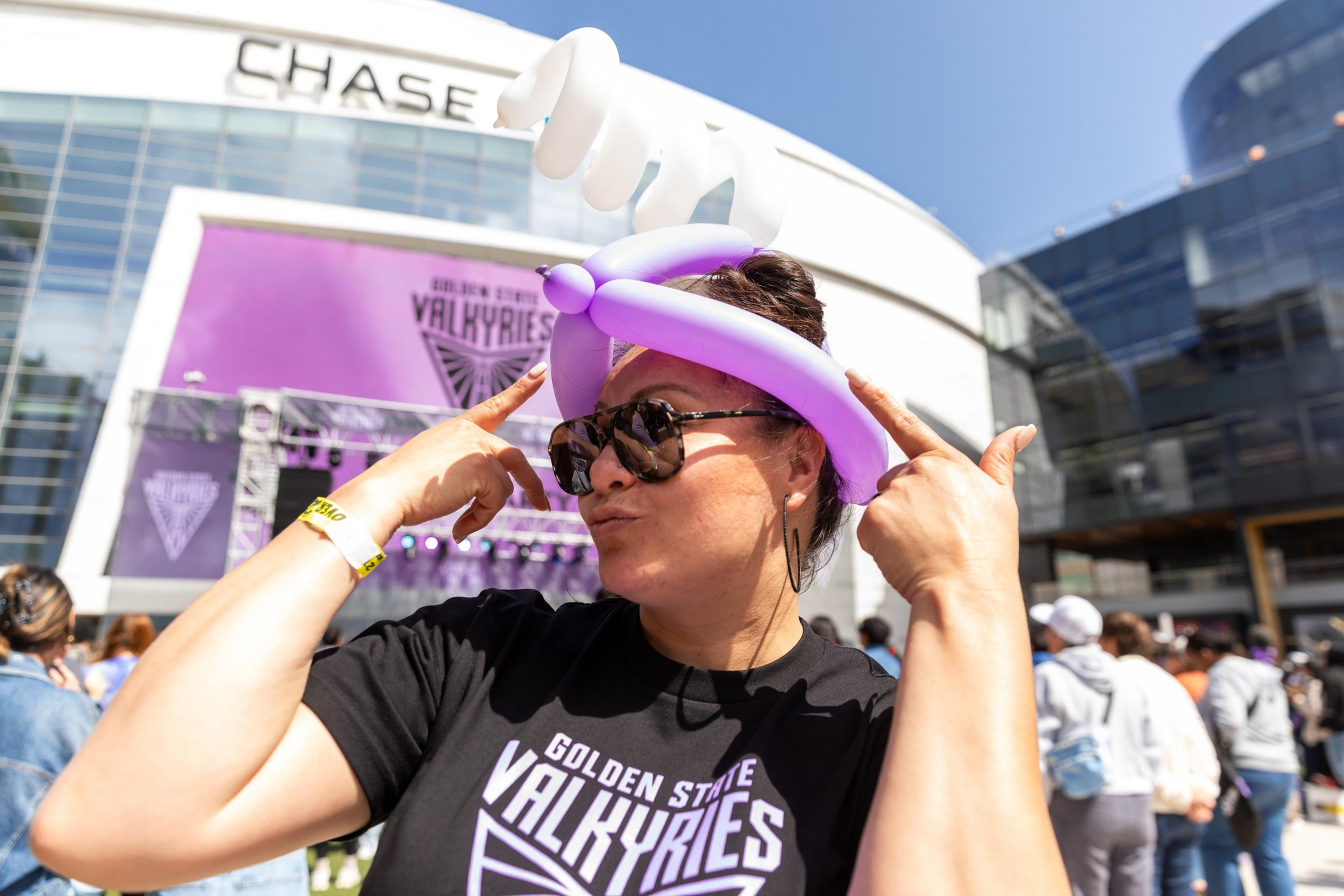 A woman gestures a peace sign, wearing sunglasses and a purple balloon hat at an outdoor event named "Golden State Valkyries" near the Chase building.