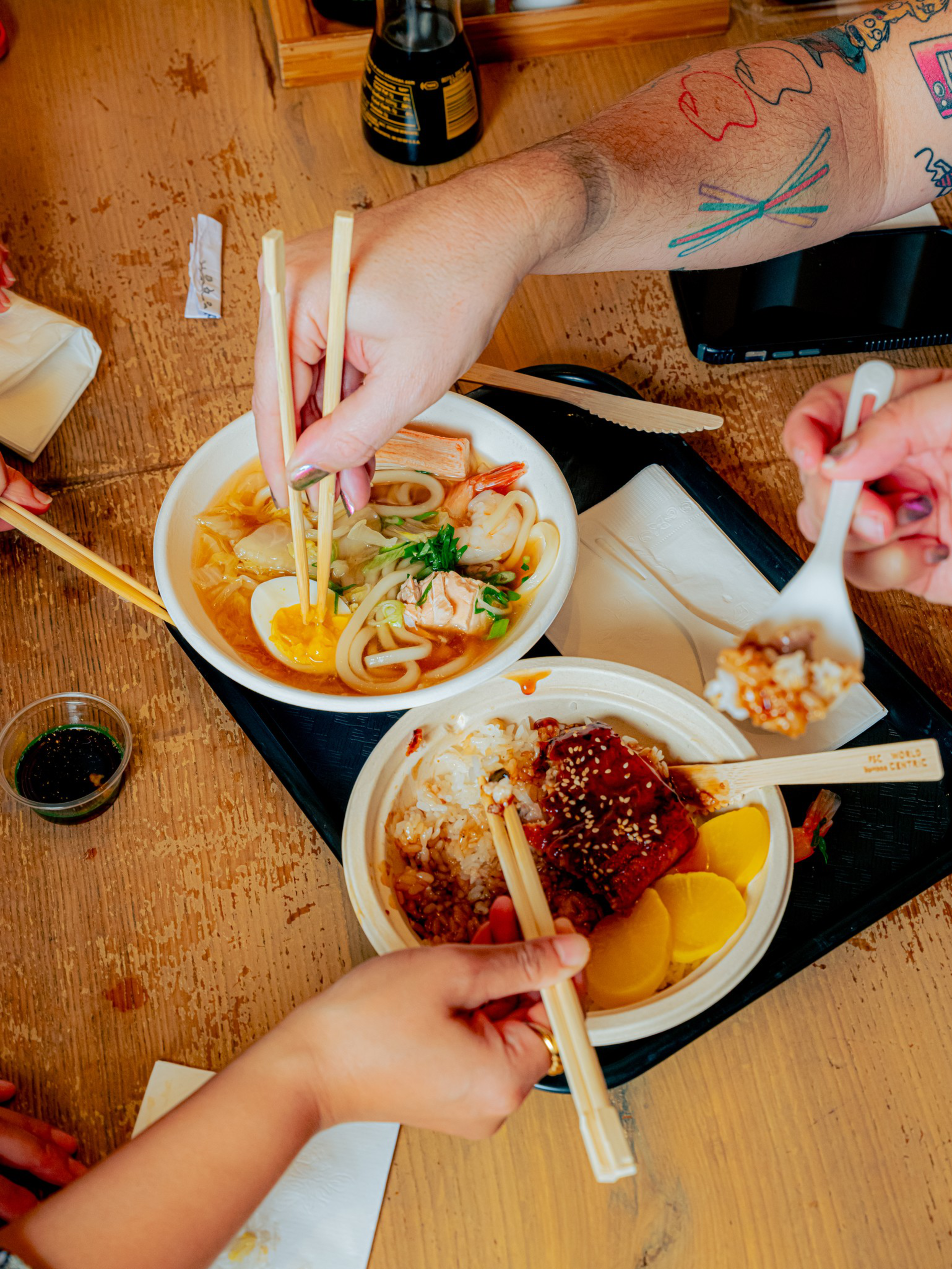 Hands with chopsticks and a spoon pick food from bowls of noodles with egg and a rice dish with meat and pickled vegetables on a wooden table.