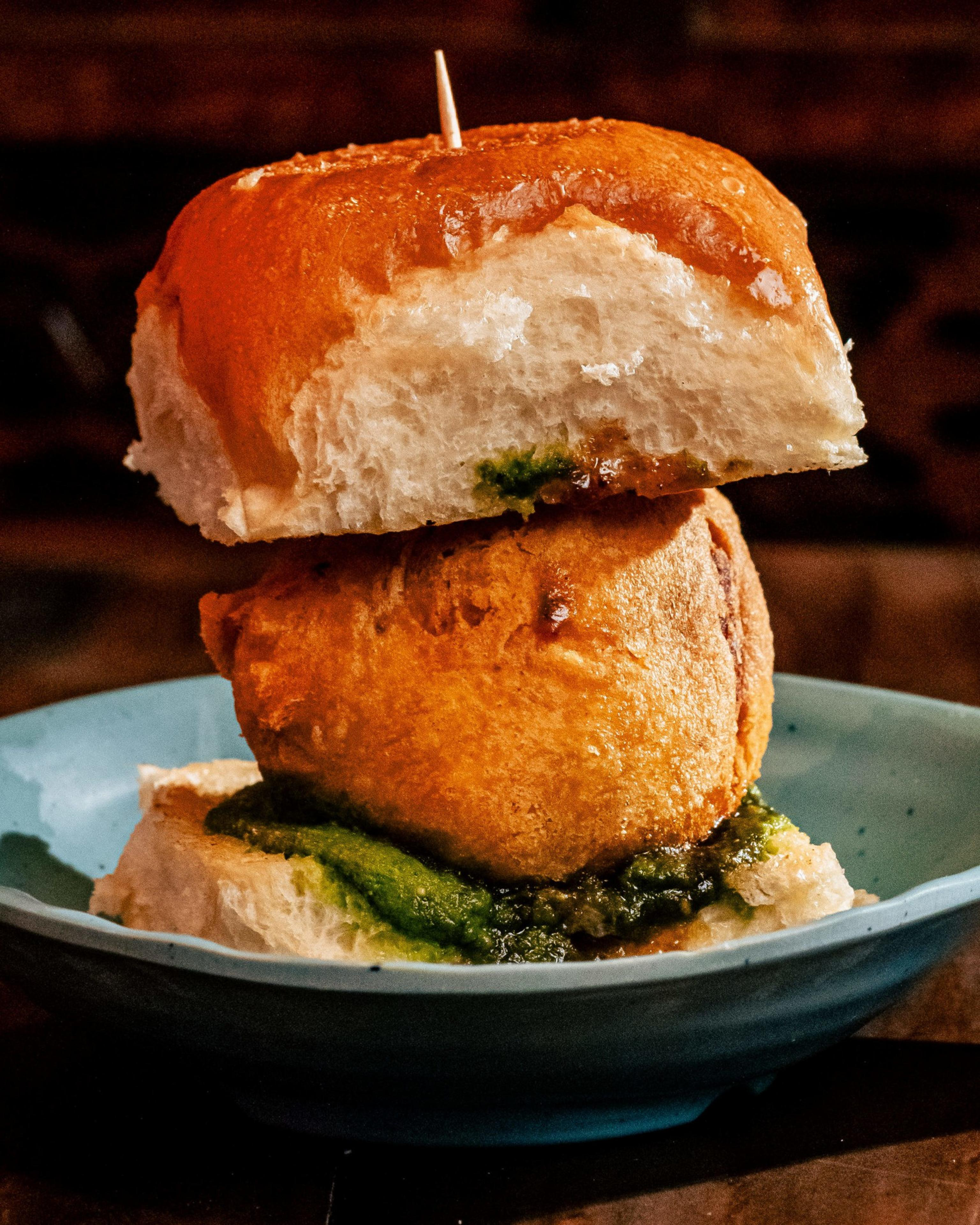 A fried ball placed between two fluffy bread slices with green chutney spread on the bottom slice, secured by a toothpick and served on a blue plate.