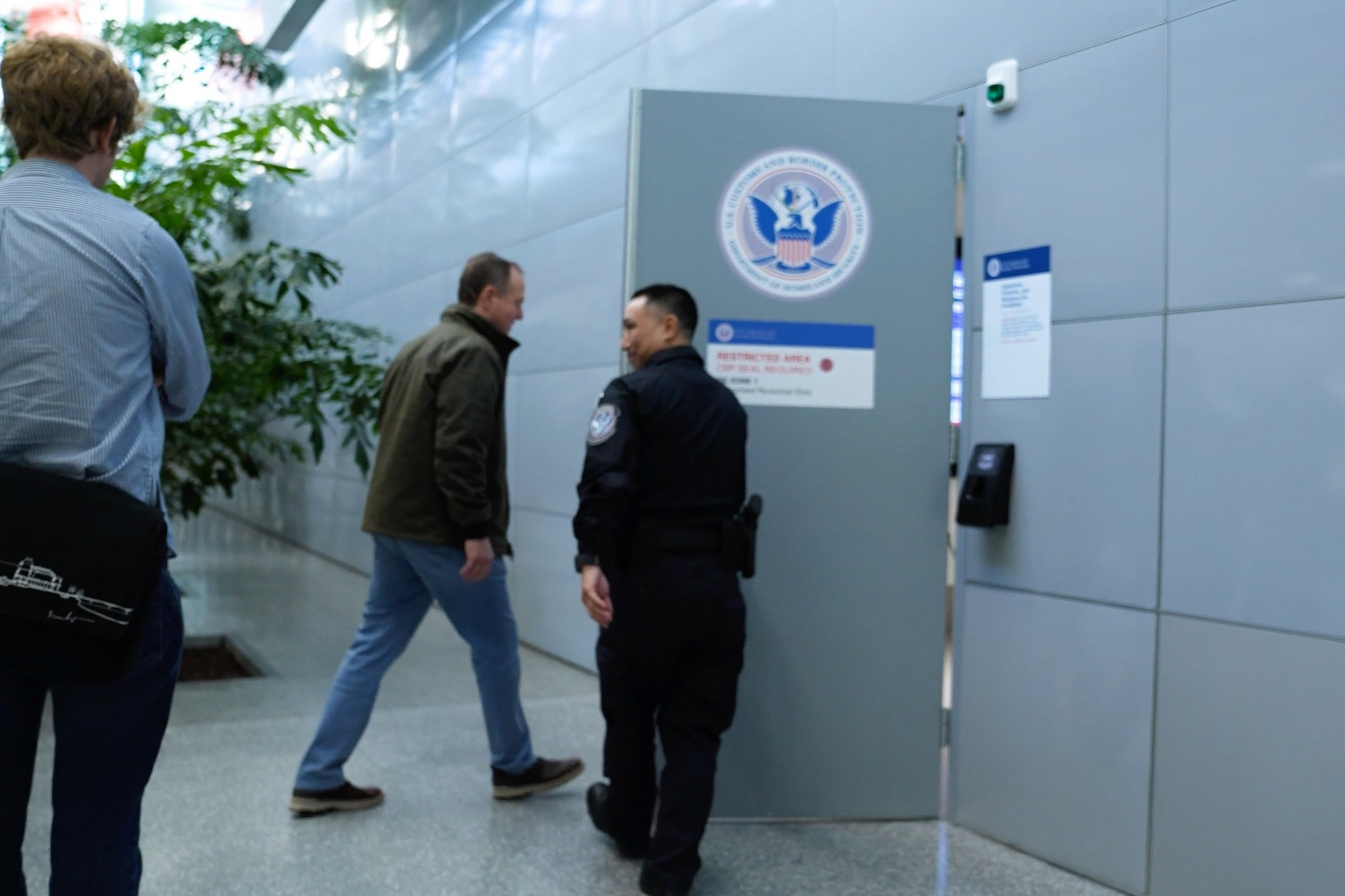 A security officer is opening a door marked with a government seal as a man walks inside and another man waits nearby.