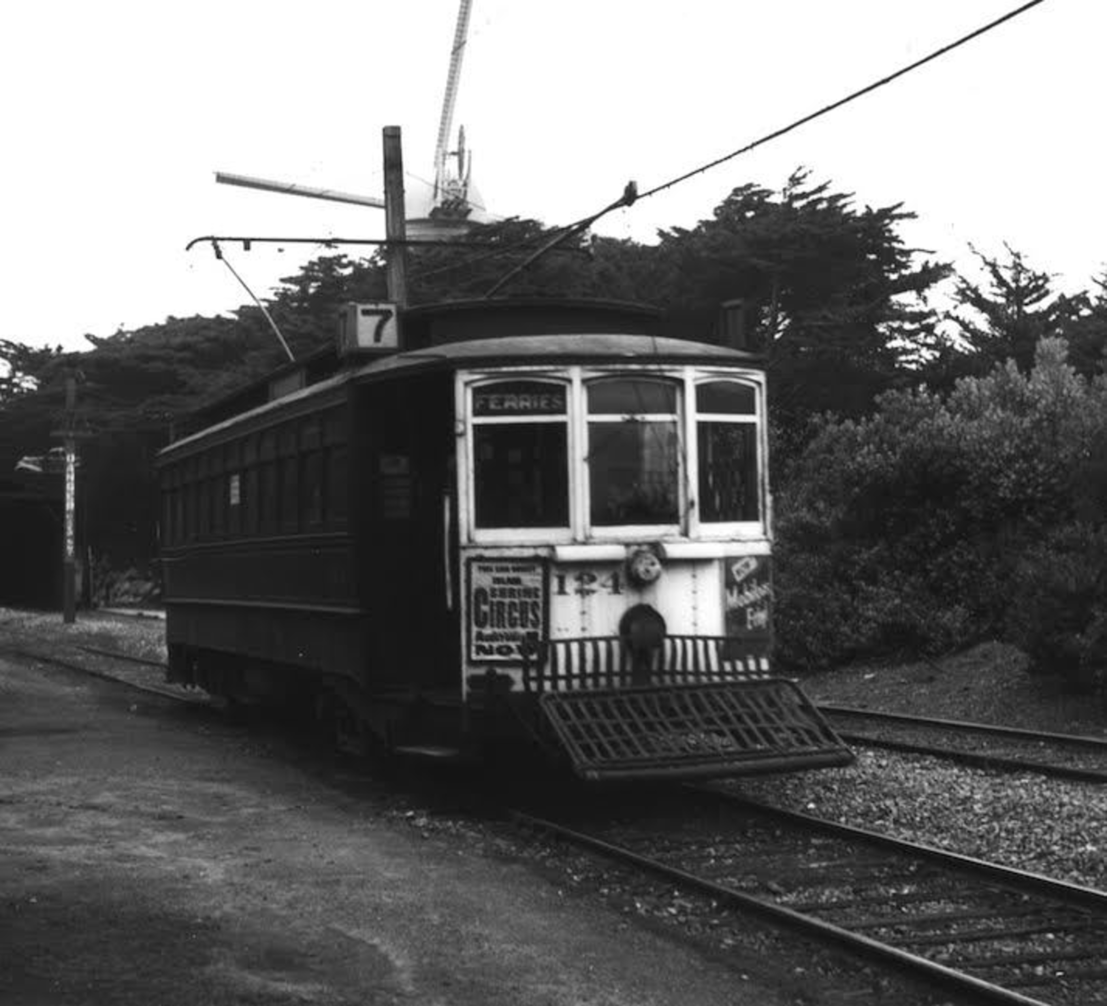 A vintage streetcar labeled "7" on tracks near trees, equipped with a front grill and advertising circus shows on its sides.