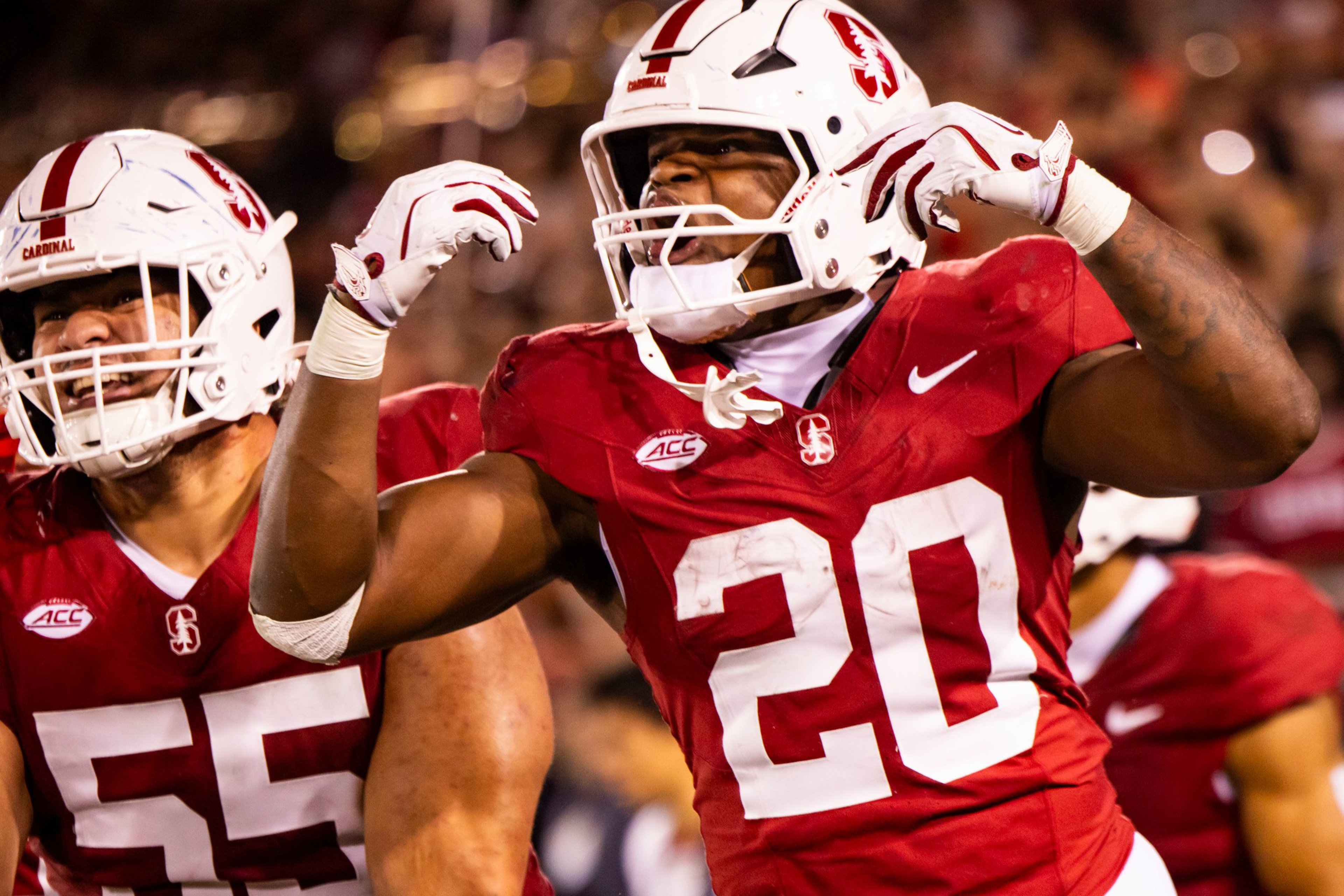 A football player wearing a red Stanford jersey with number 20 and a white helmet celebrates by flexing his arms, while a teammate in jersey 55 smiles nearby.