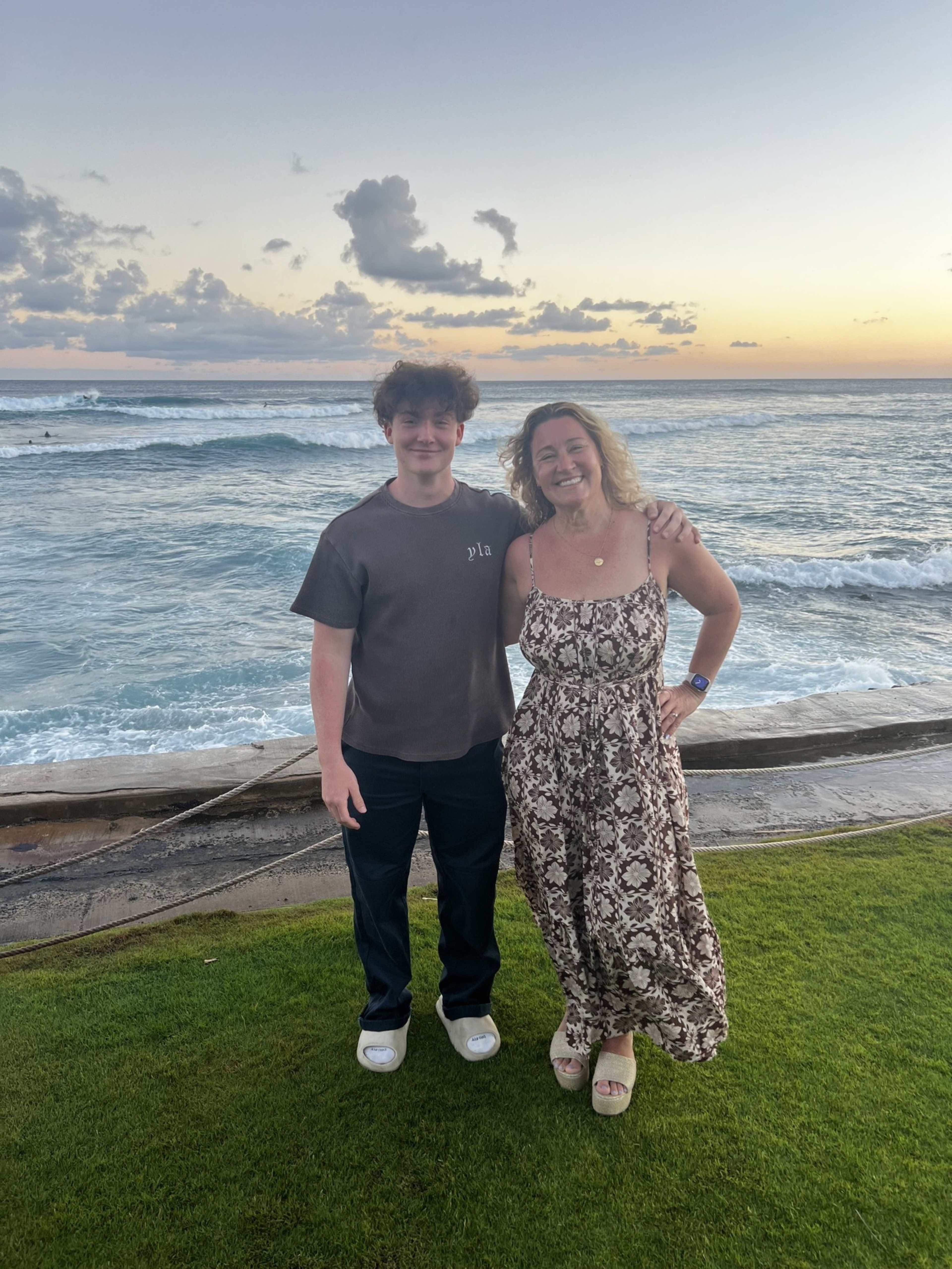 A smiling young man and woman stand side by side on grass near the ocean at sunset, with the woman’s arm around the man’s shoulder.