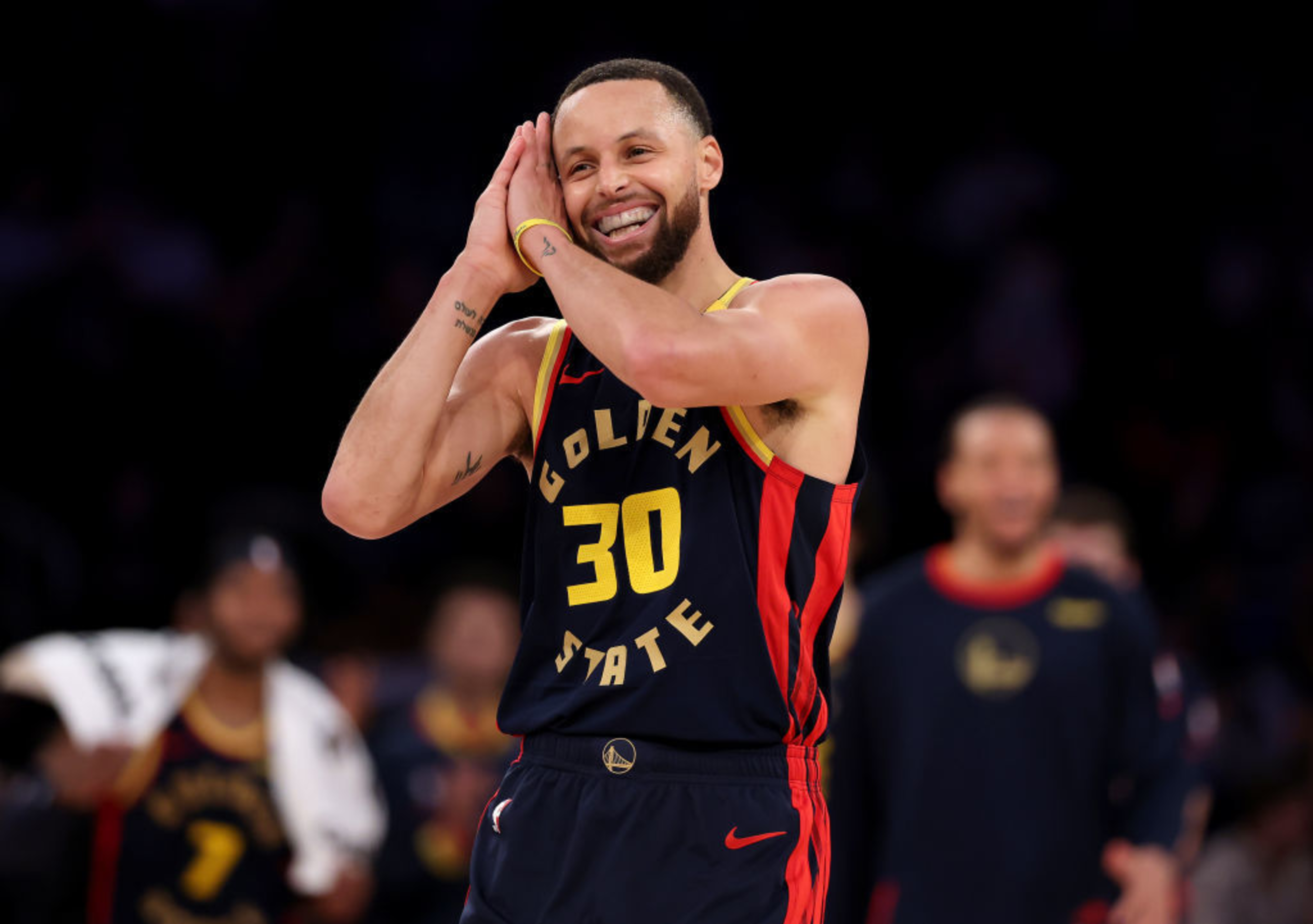 A basketball player in a navy and gold jersey numbered 30 is smiling and mimicking a sleep gesture with his hands on his cheek. Others are blurred in the background.