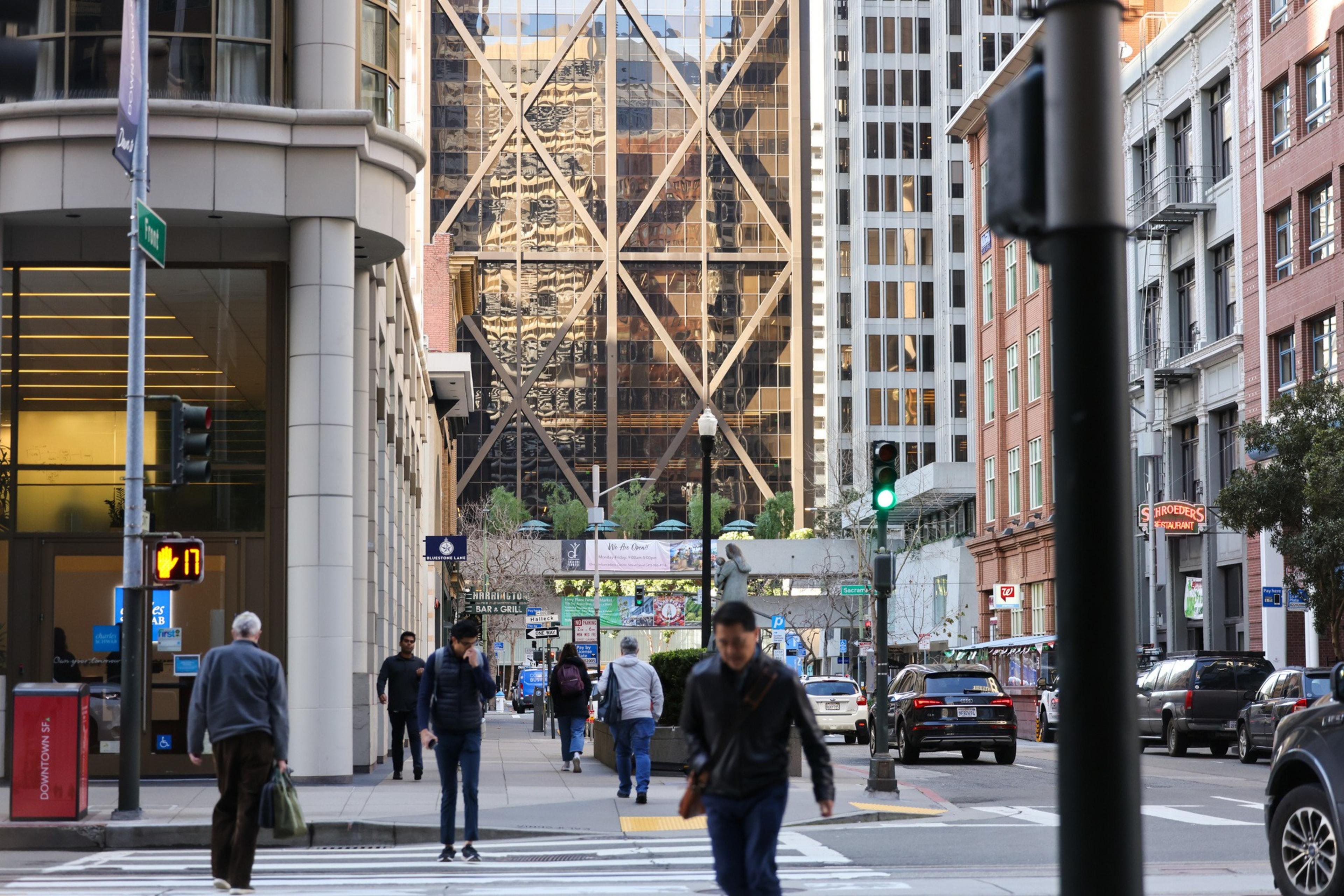 People walk along a busy city street, surrounded by tall buildings and a reflective, geometric-patterned skyscraper. Traffic signals and shop signs are visible.