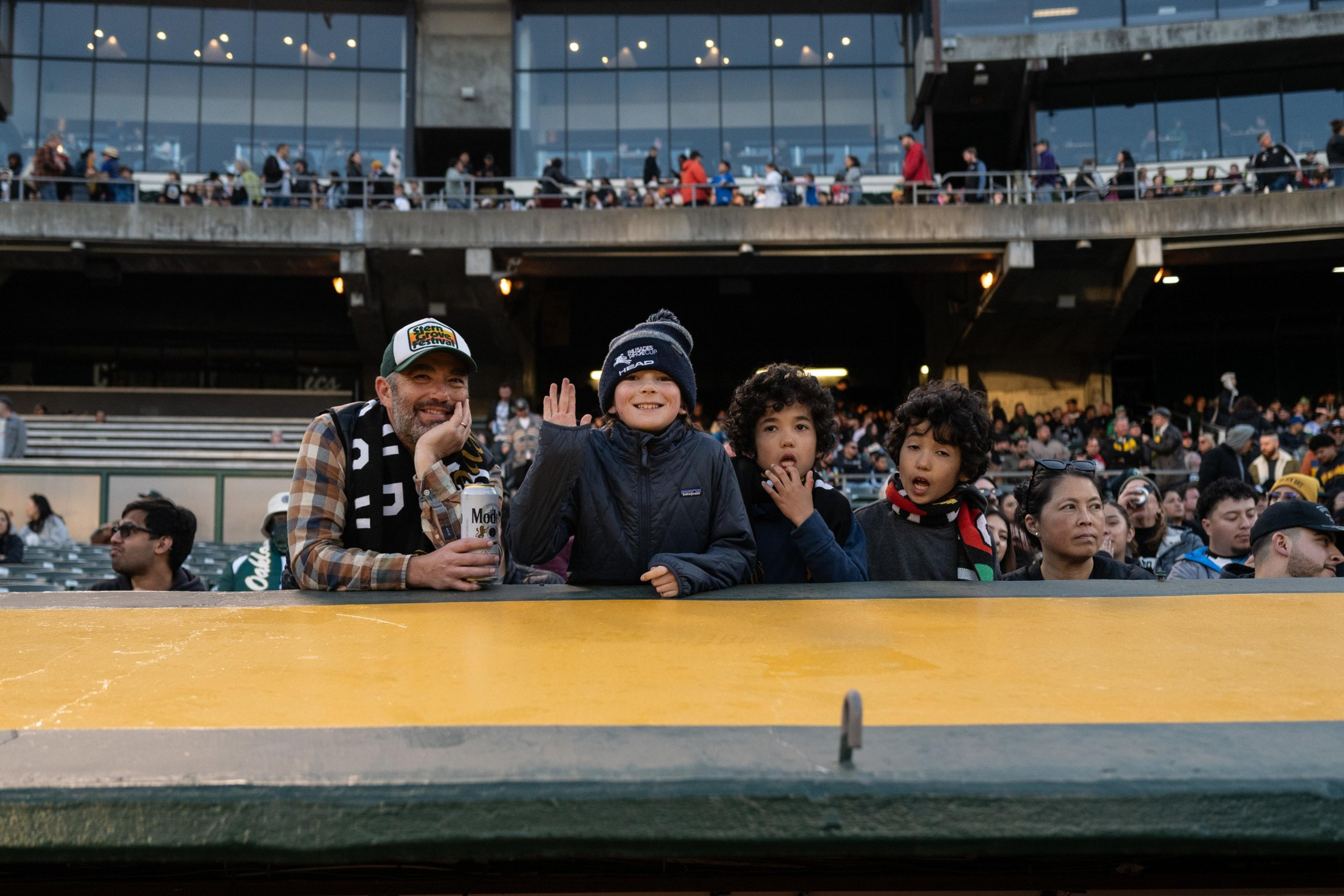 A man and four children are sitting in a stadium. The man smiles and holds a drink, while one child waves. The background shows a full audience.