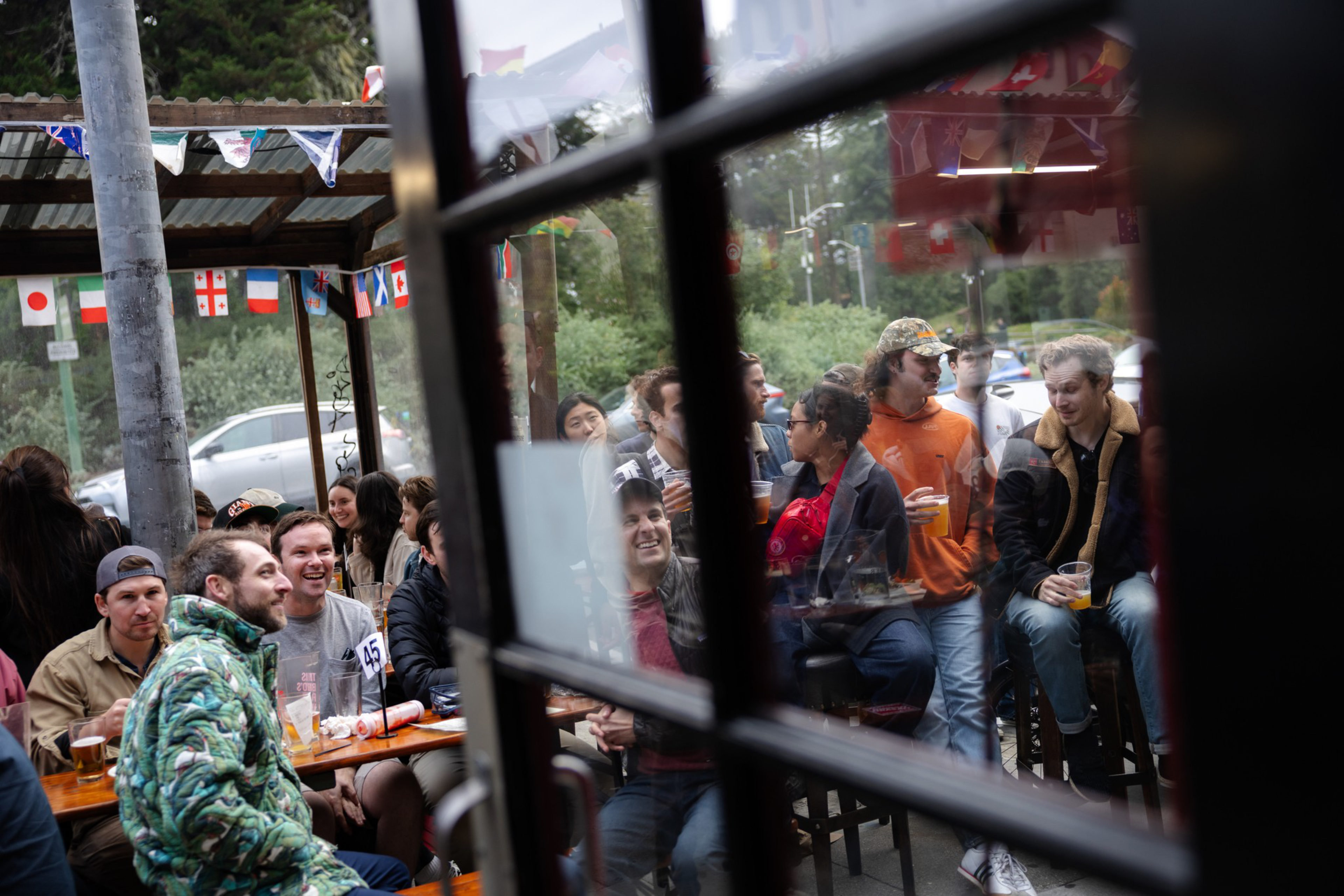 People sit and stand closely together at an outdoor pub or cafe, chatting and enjoying drinks under a roof decorated with international flags.
