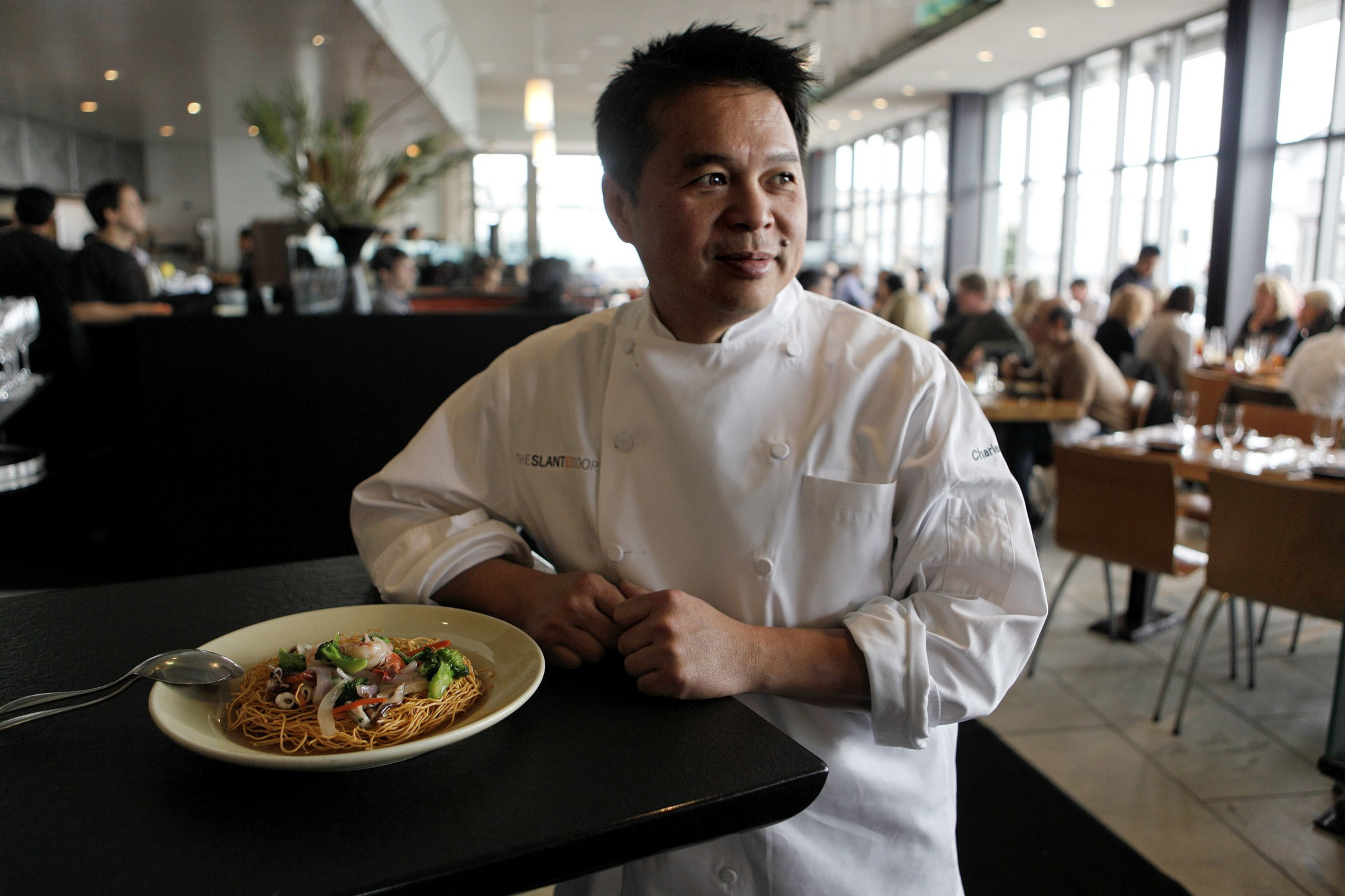 A chef in a white uniform stands beside a plate of noodles garnished with vegetables, in a busy restaurant with people dining in the background.