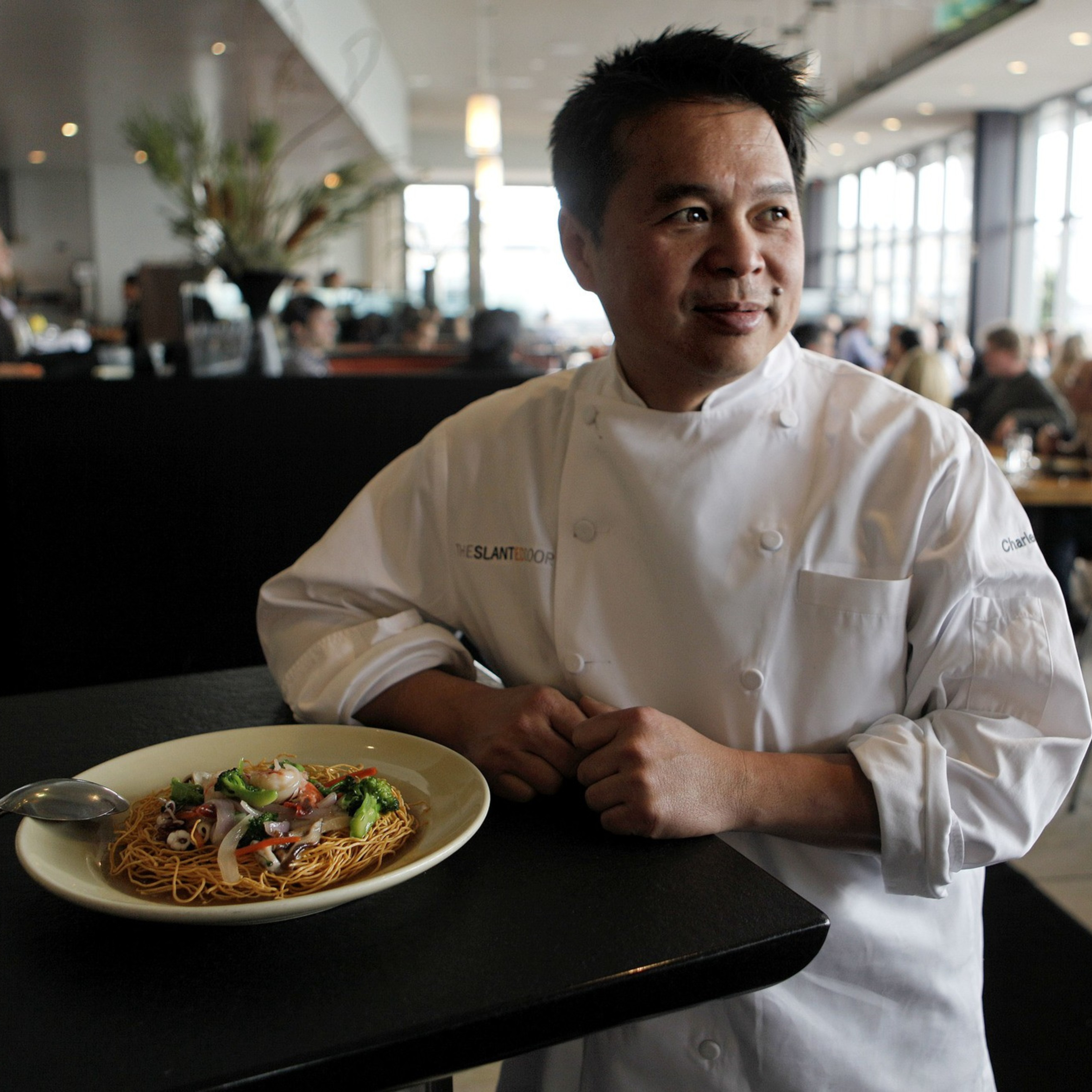 A chef in a white uniform stands beside a plate of noodles garnished with vegetables, in a busy restaurant with people dining in the background.