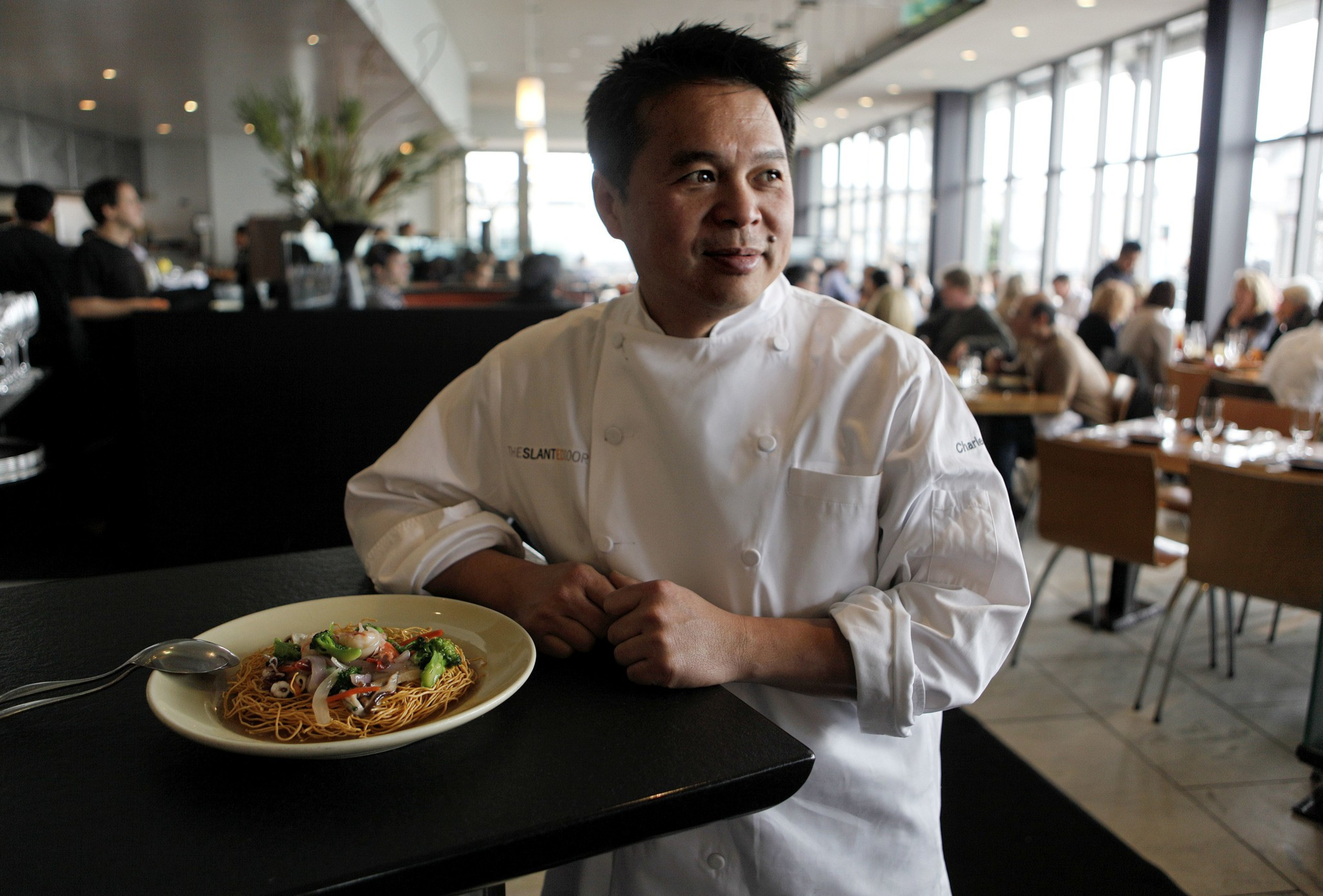 A chef in a white uniform stands beside a plate of noodles garnished with vegetables, in a busy restaurant with people dining in the background.