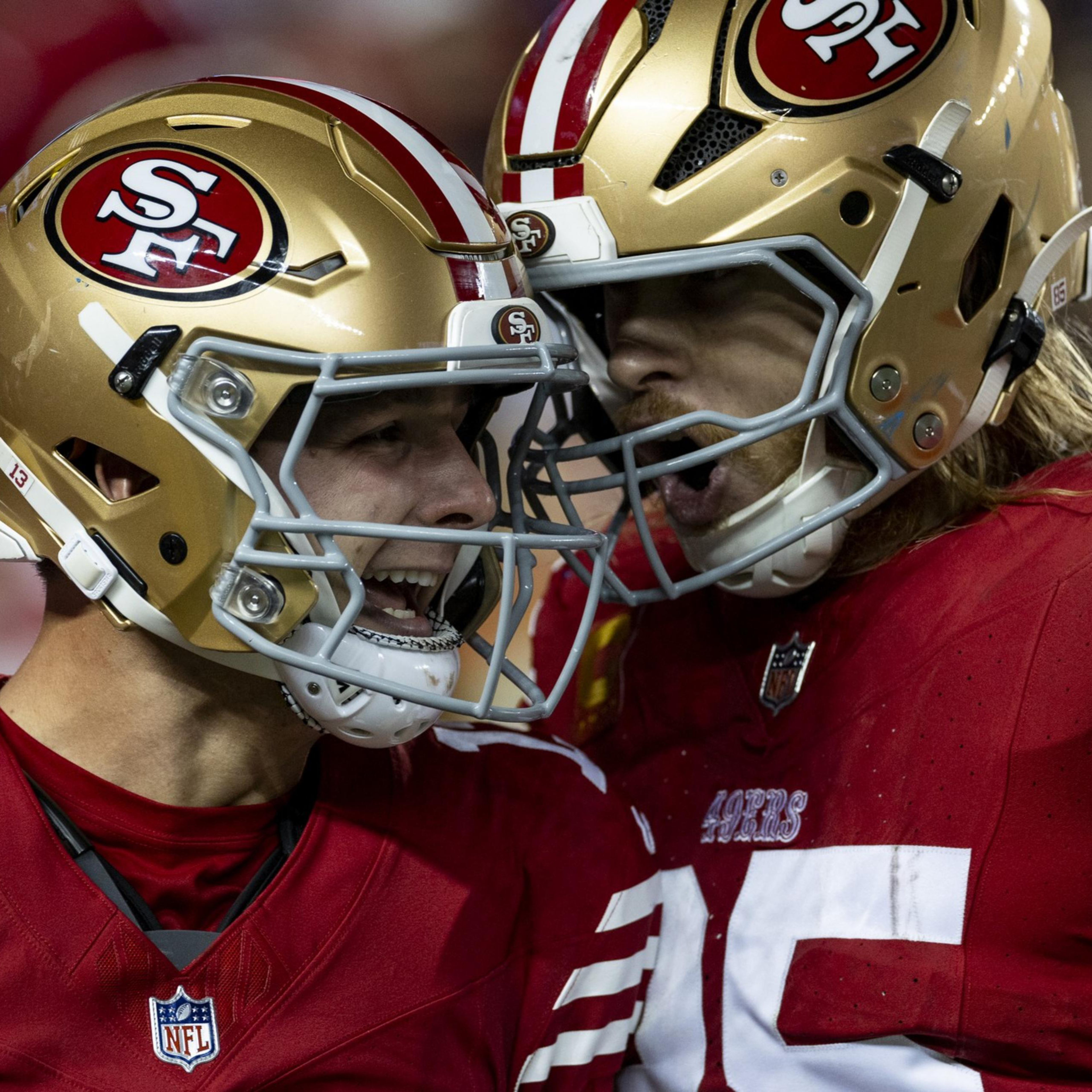 Two football players wearing red uniforms and gold helmets with the 49ers logo are celebrating on the field, their expressions are enthusiastic and intense.
