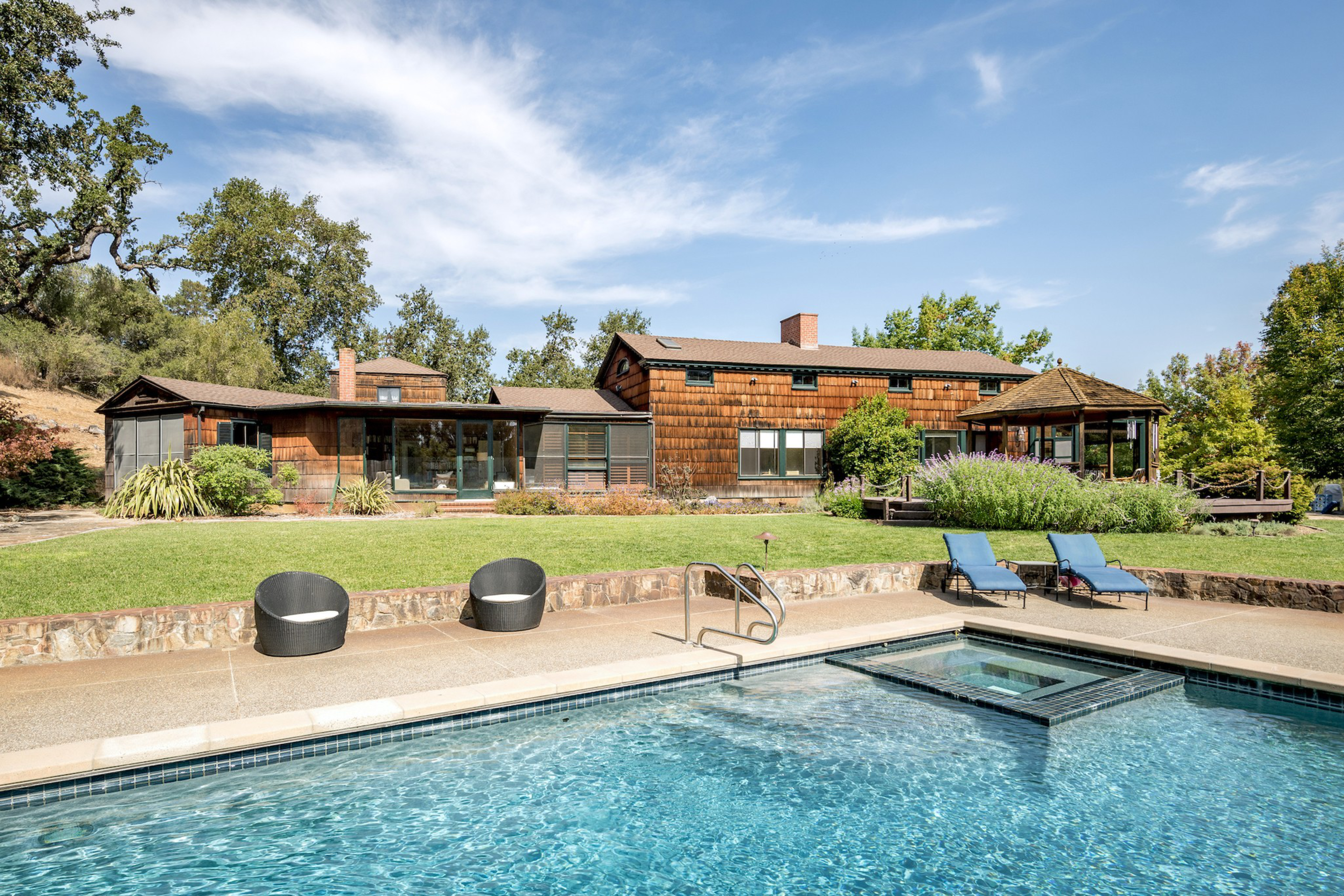 A backyard with a swimming pool, two black round chairs, two blue lounge chairs, a wooden house, green lawn, and trees under a blue sky.