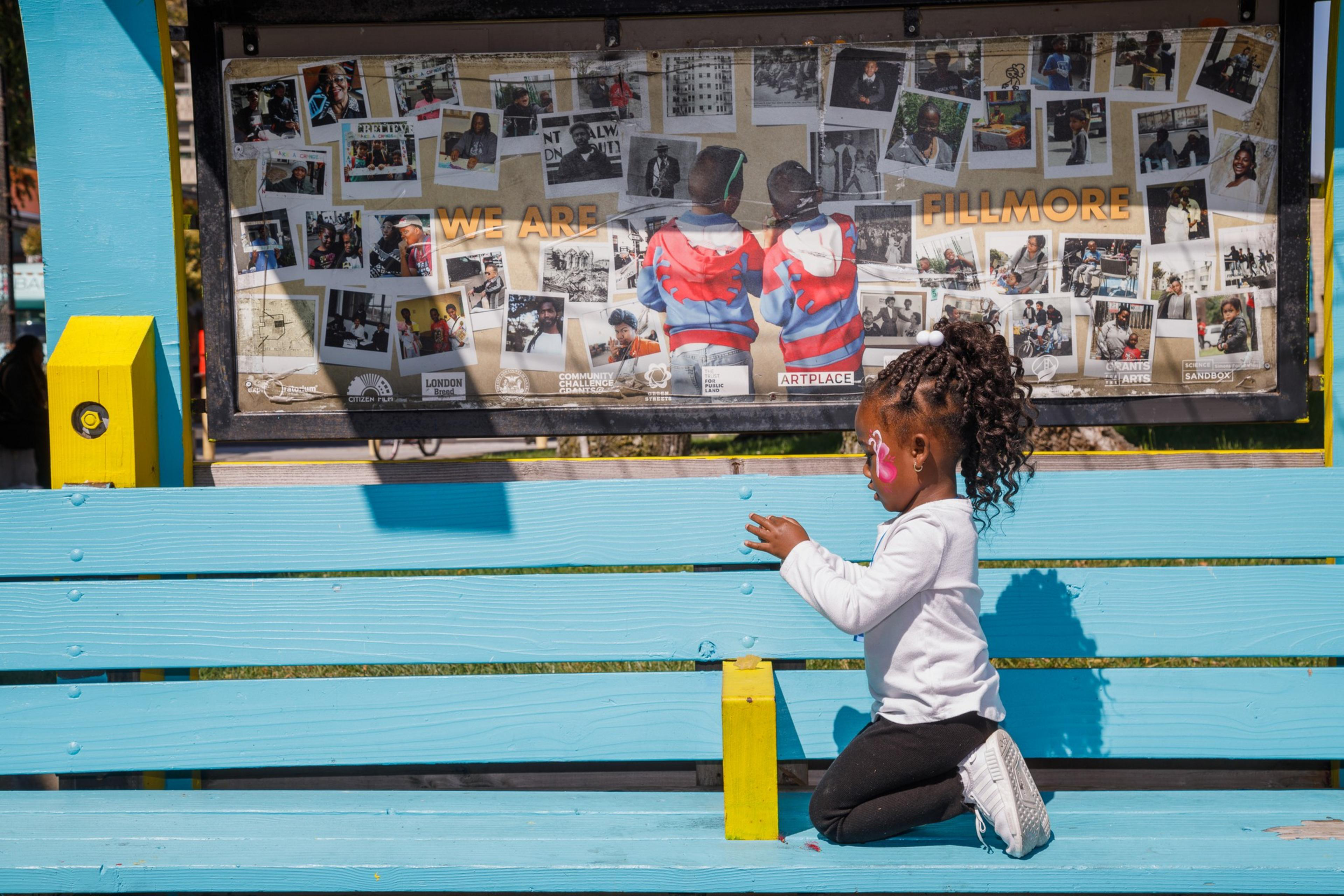 A child sits on a vibrant blue bench, facing a collage of photos on a bulletin board, under text &quot;WE ARE FILLMORE&quot;.