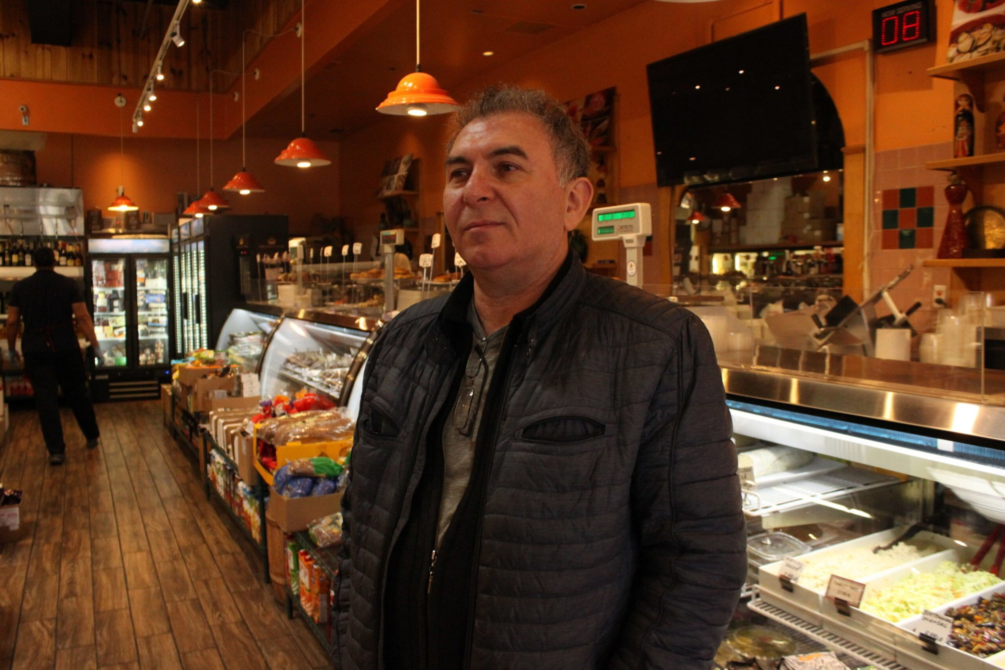 A man in a dark jacket stands inside a warmly lit store with wooden floors, surrounded by refrigerated displays and shelves stocked with food.