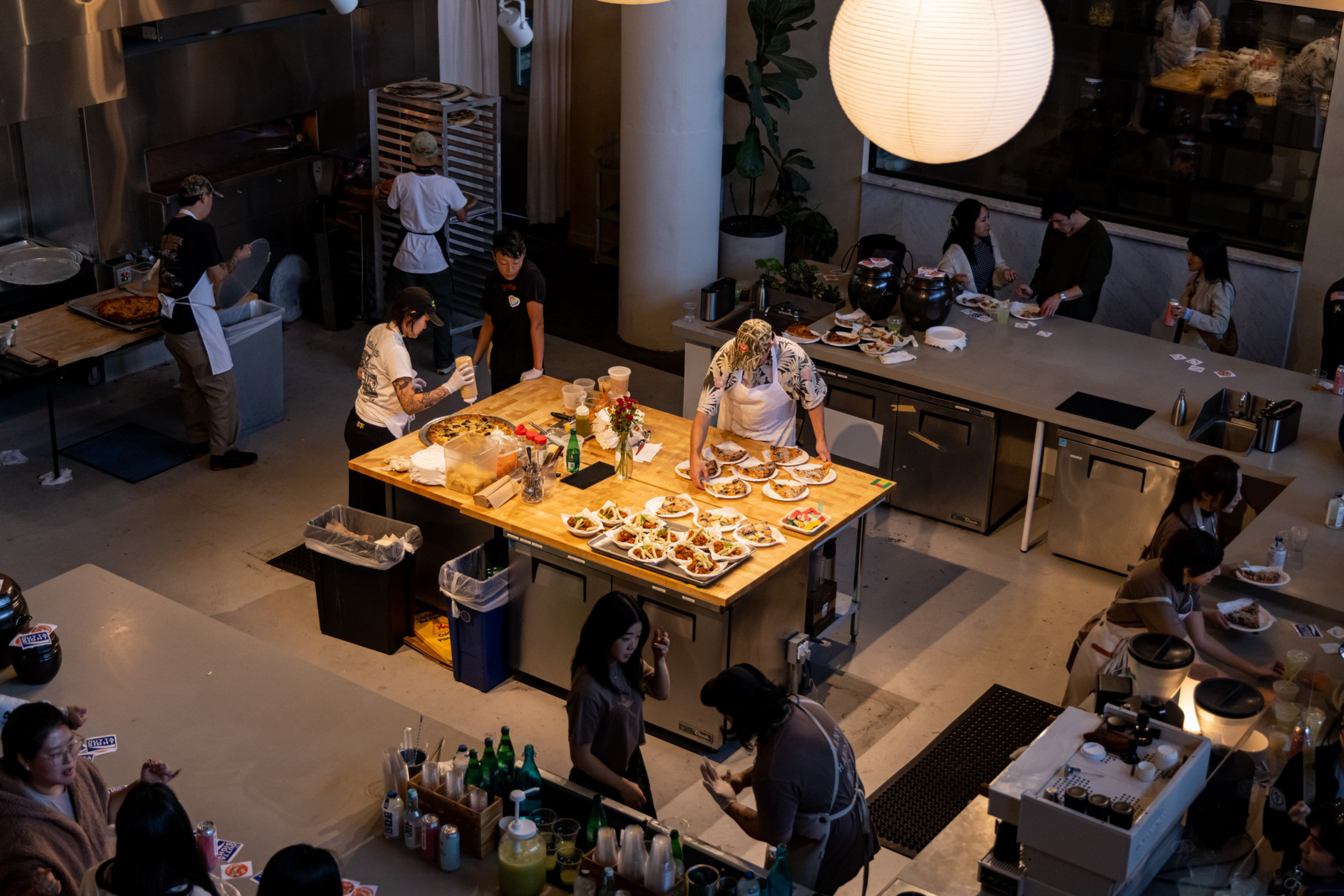 A busy kitchen with staff preparing and plating food on a central wooden table, while others work or eat nearby under warm lighting.