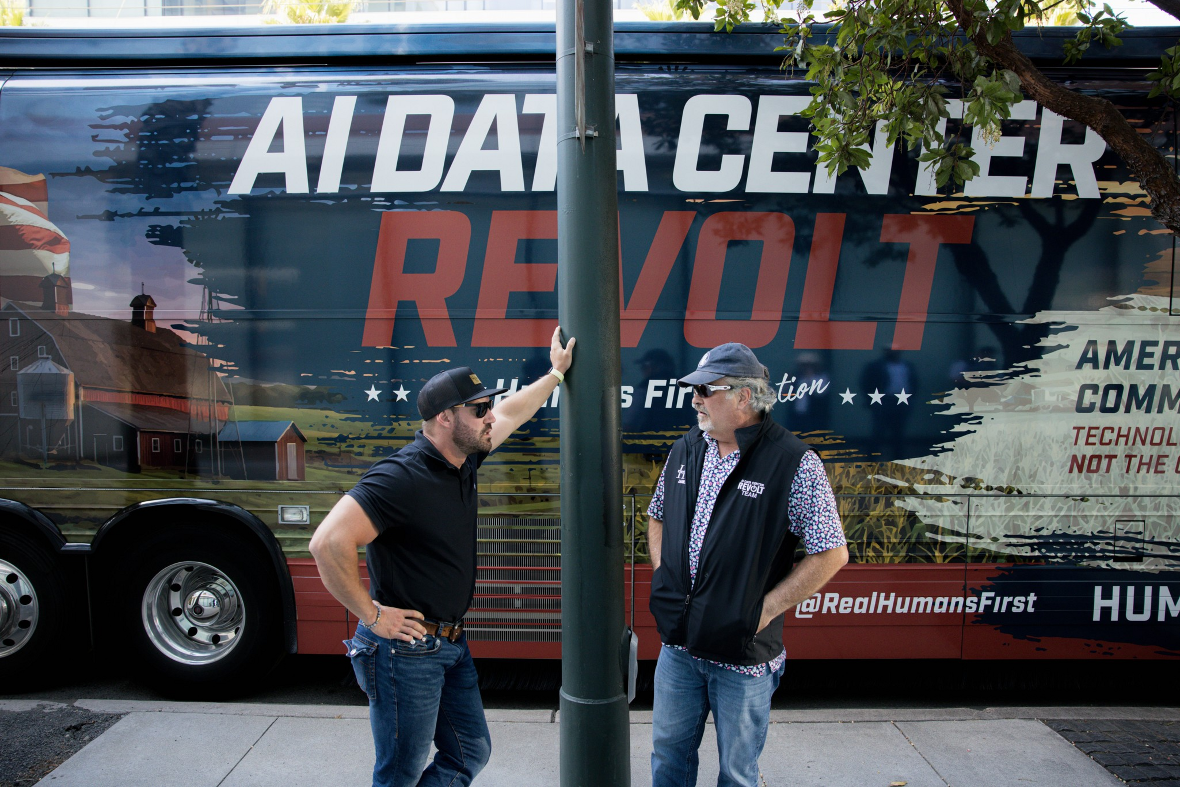 Two men talk on a sidewalk beside a large bus with text reading "AI Data Center Revolt" and "Humans First" on its side.