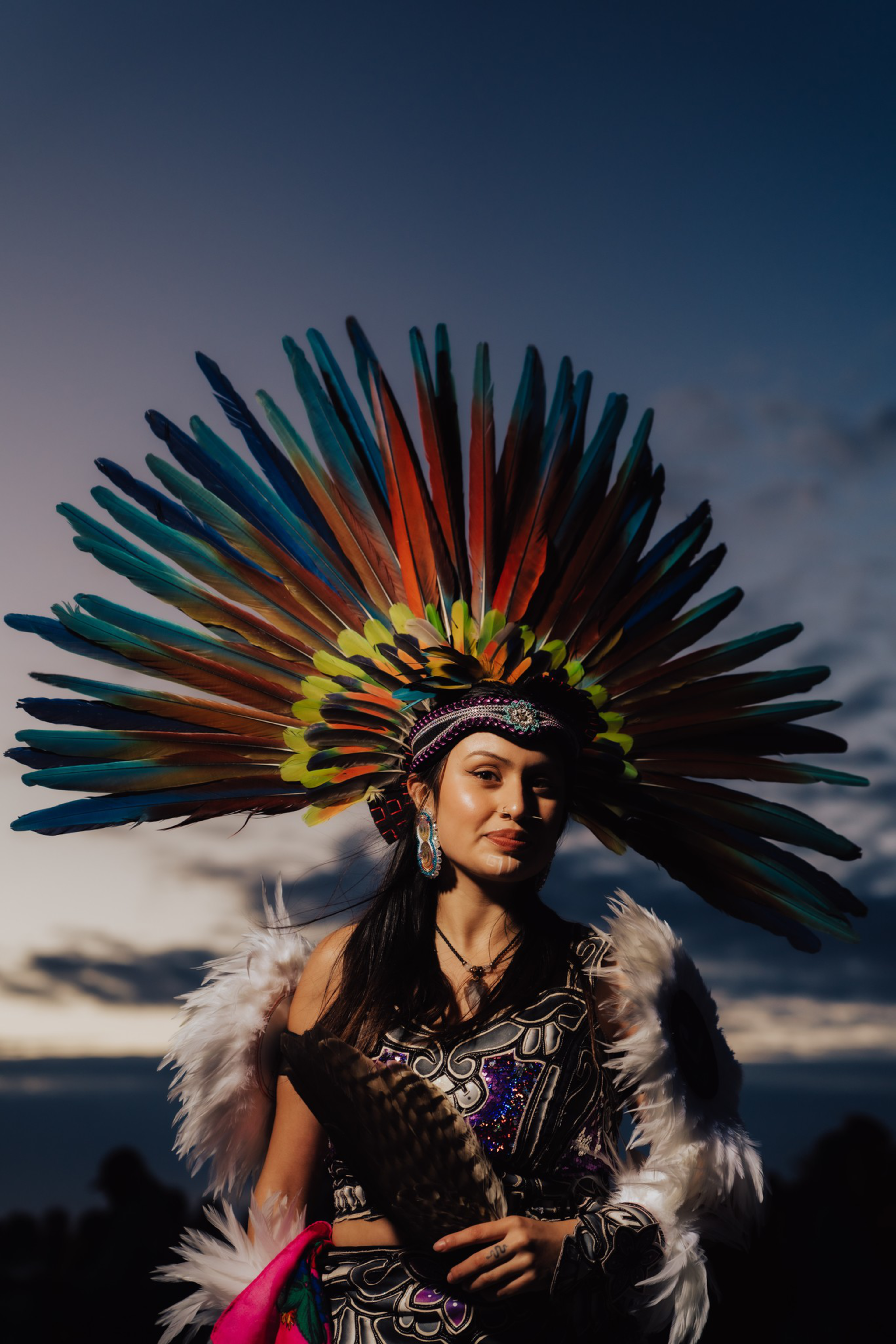 A woman wears a colorful feathered headdress and traditional attire with feathered arm bands, holding a fan, against a dusk sky background.