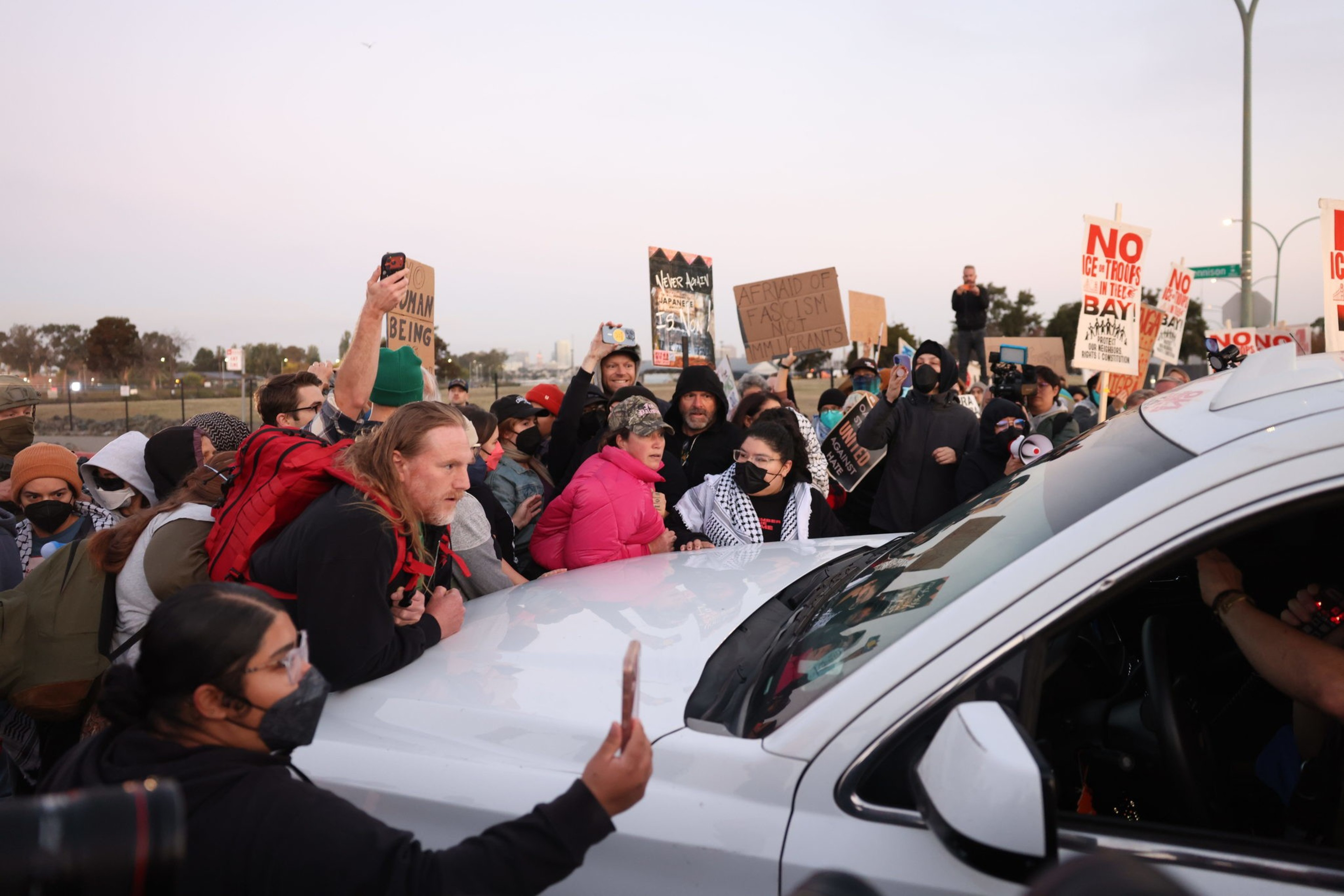 A crowd of protesters surrounds a white vehicle, holding signs against ICE and fascism while recording the scene on their phones.