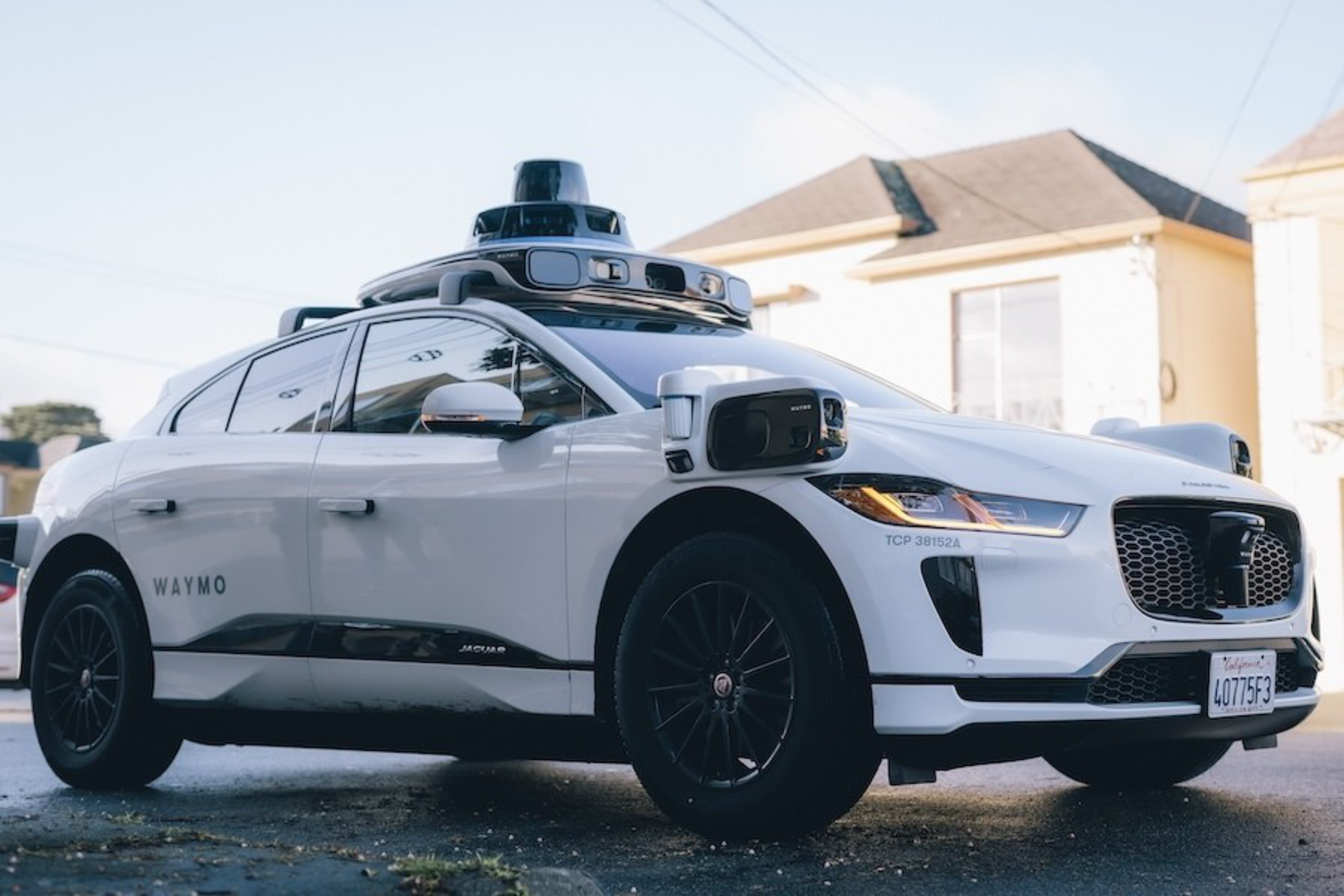 A white self-driving Jaguar I-PACE with &quot;WAYMO&quot; branding and equipped with sensors on a sunny street.