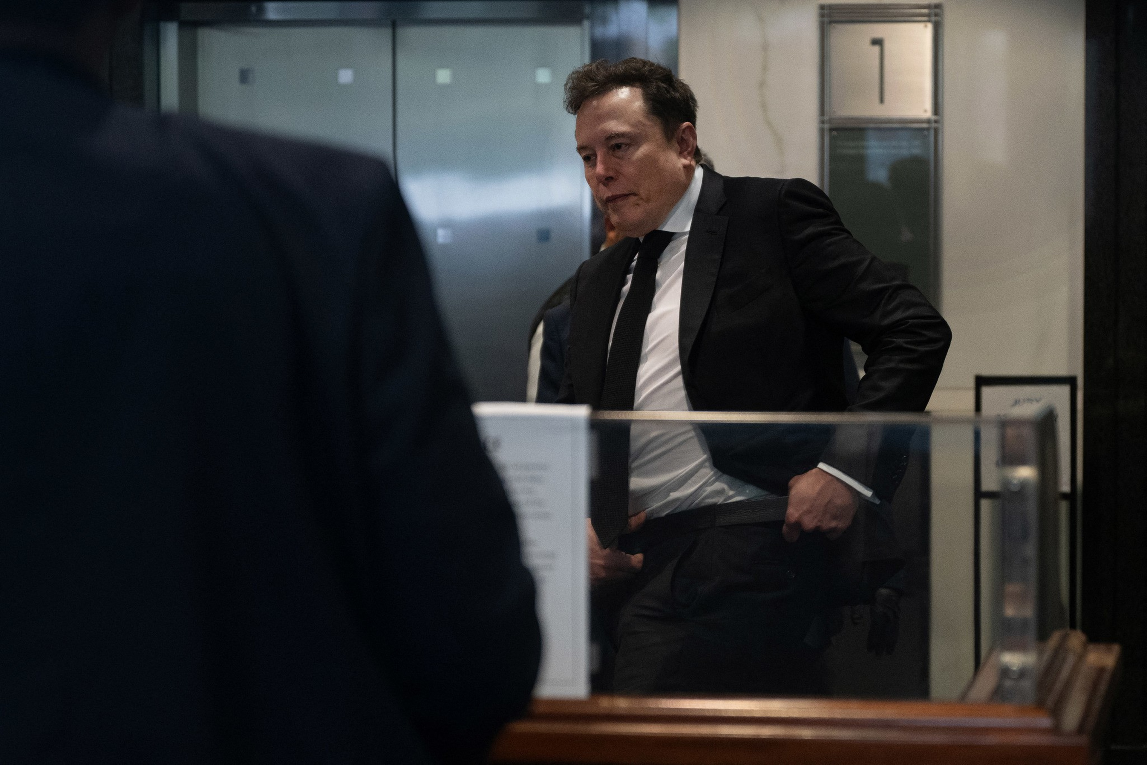 A man in a black suit adjusts his jacket while standing behind a glass barrier, with an elevator and a number “1" sign in the background.
