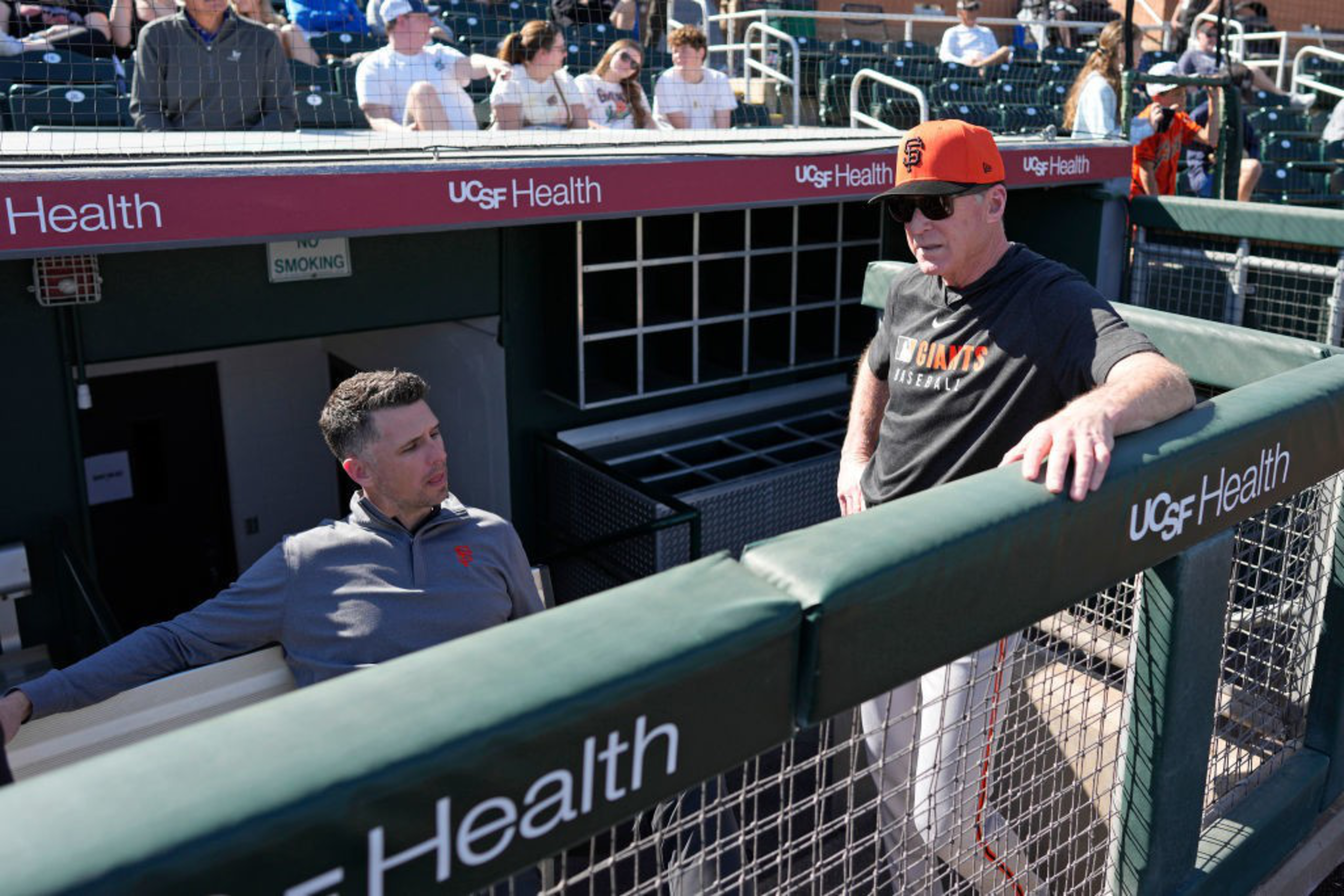Two men in a dugout are talking casually. One is seated, wearing a gray jacket, and the other, standing, wears a black "Giants Baseball" shirt and cap.